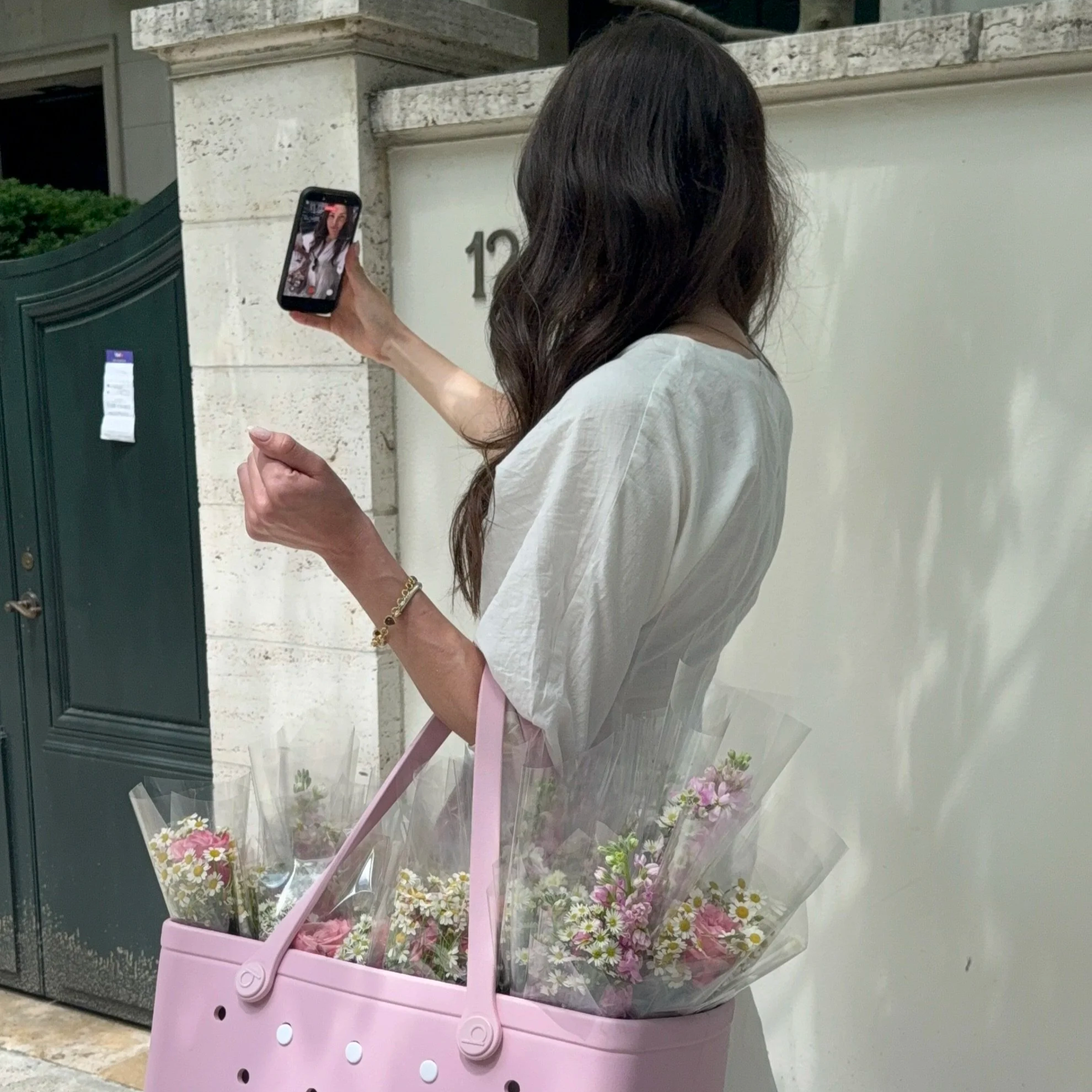 A woman with long dark hair taking a selfie with her phone while standing next to a decorative flower cart filled with pink and white flowers outside a building.