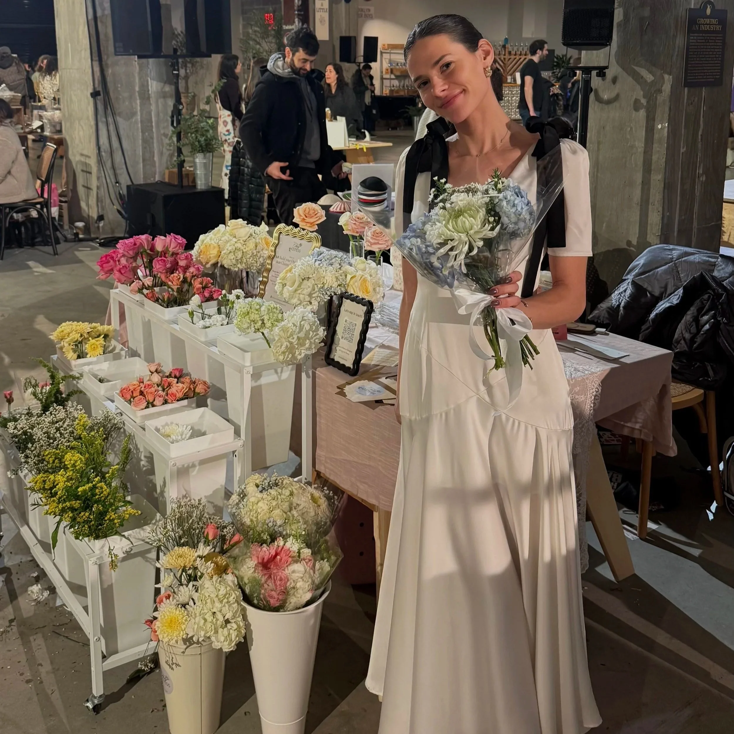 A woman with dark hair tied back, wearing earrings and a white dress, holding a bouquet of flowers, smiling at a floral display in a store or market setting with other people and floral arrangements around.