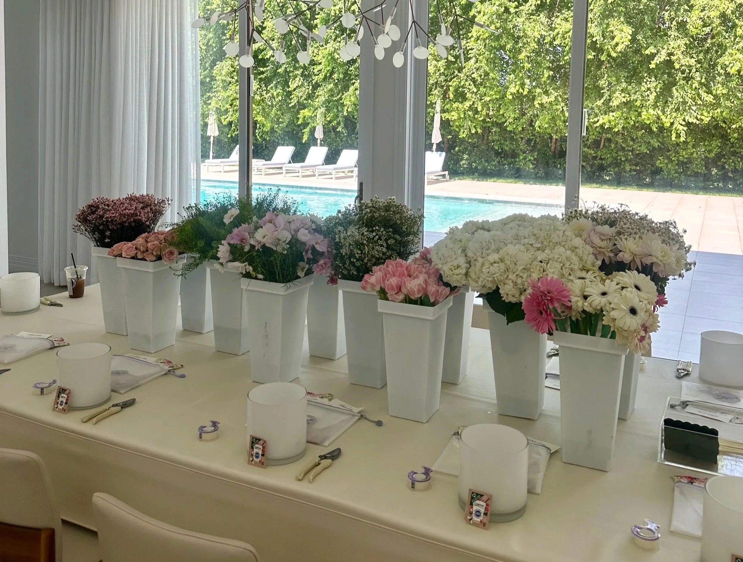 A table decorated with white vases filled with various pink and white flowers, set near a large window overlooking a pool and trees outside.