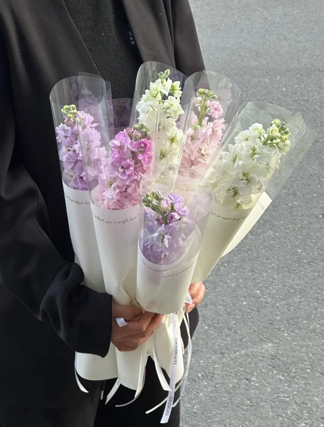 A person holding five bouquets of pastel-colored flowers wrapped in white paper and transparent plastic, standing on a paved surface.