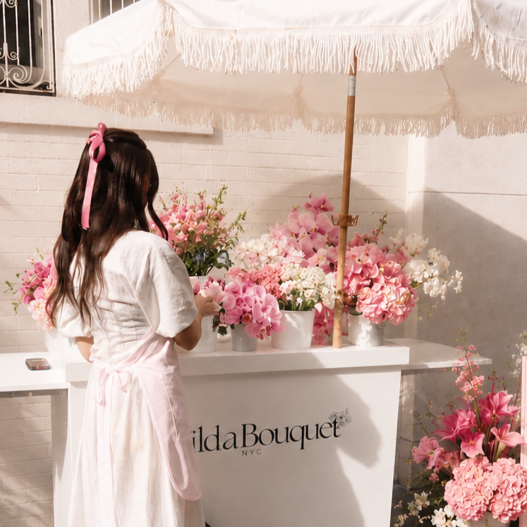 A woman with long brown hair tied with pink ribbon, wearing a white dress and pink apron, arranging pink and white flowers at a floral stand under a white umbrella.