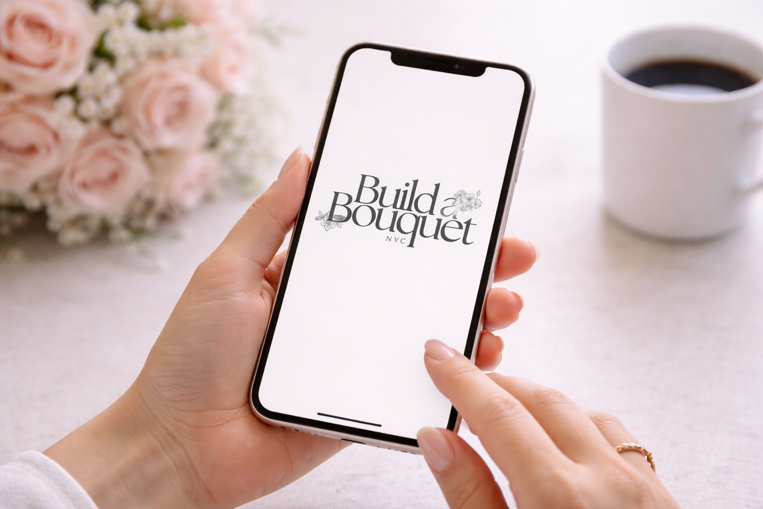 Person holding a smartphone with 'Build a Bouquet NYC' displayed on the screen, a bouquet of pink roses and white flowers in the background, and a cup of coffee on a light-colored table.