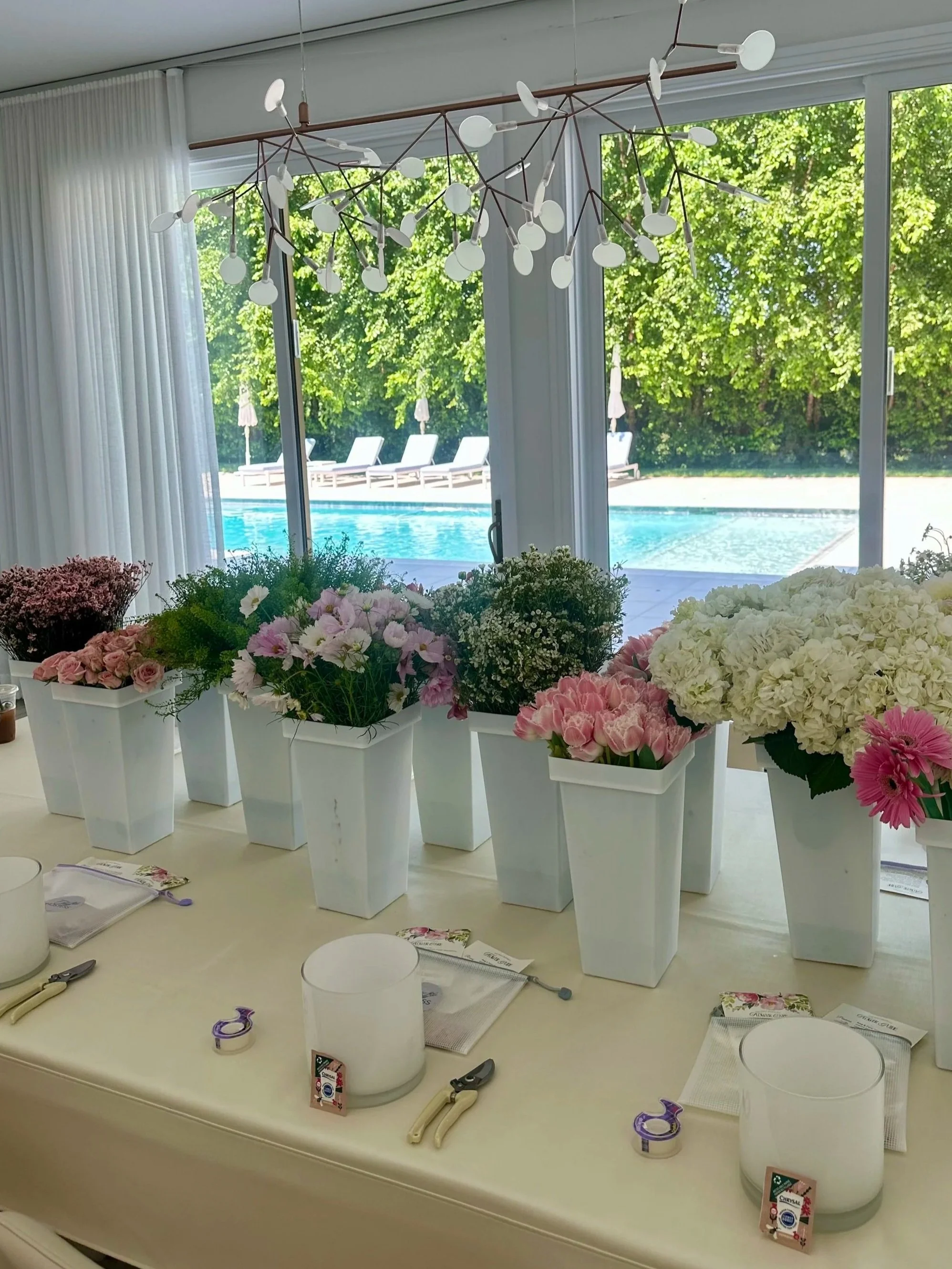 A room with large windows showing green trees and a swimming pool outside, decorated with a modern chandelier and a table with various flowers and gardening supplies, including pots, scissors, and plant labels.