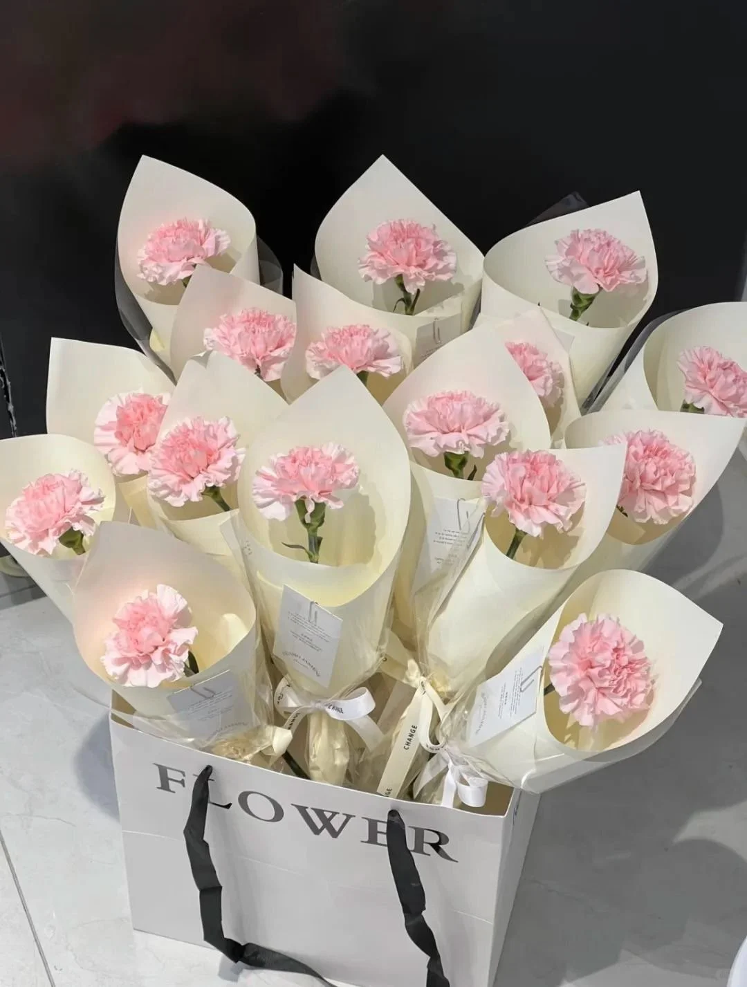 A white shopping bag labeled 'FLOWER' filled with multiple pink carnations in white wrapping paper.