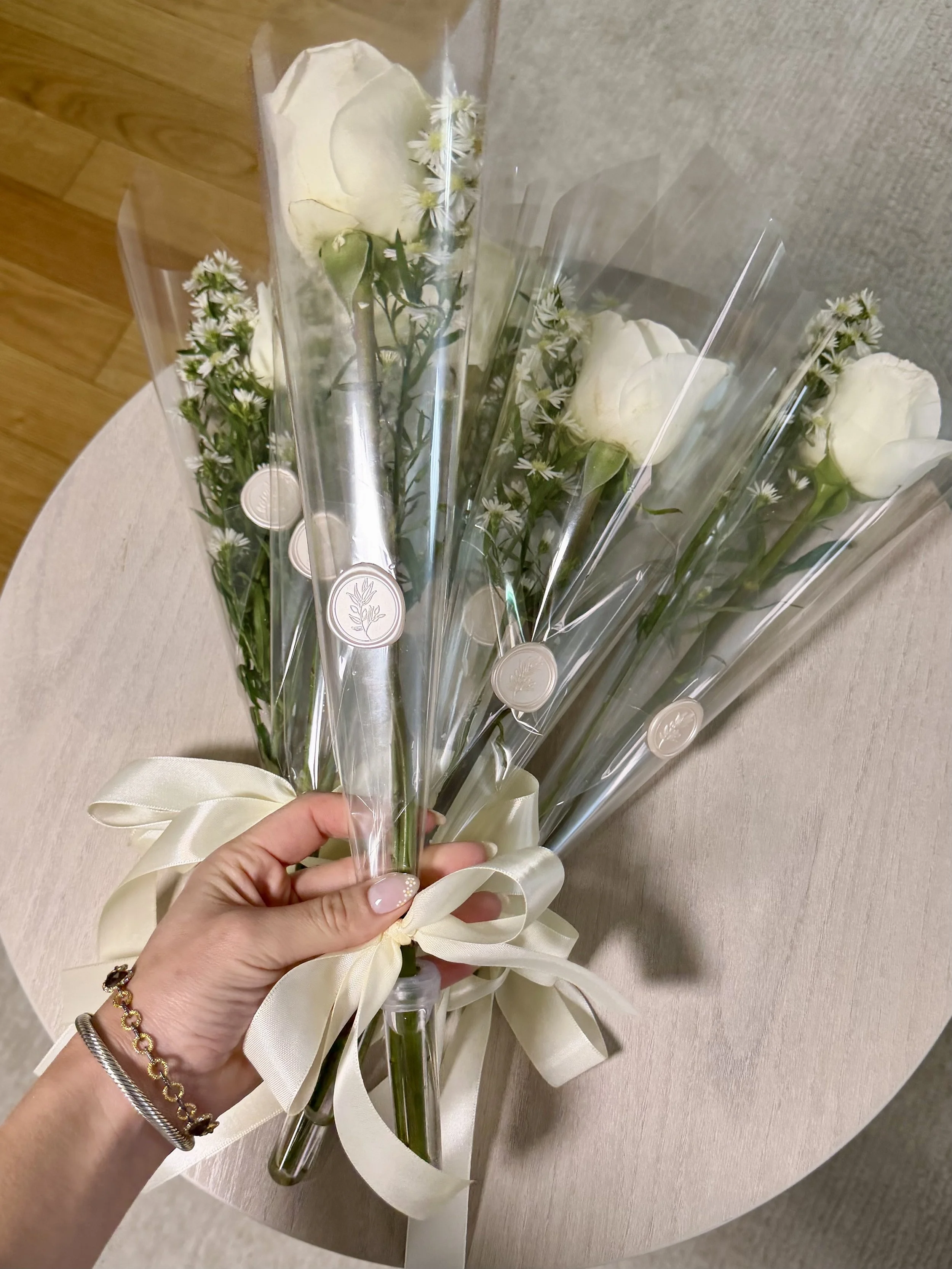 A bouquet of white roses and small white flowers wrapped in clear plastic with a cream-colored ribbon, held by a person's hand.