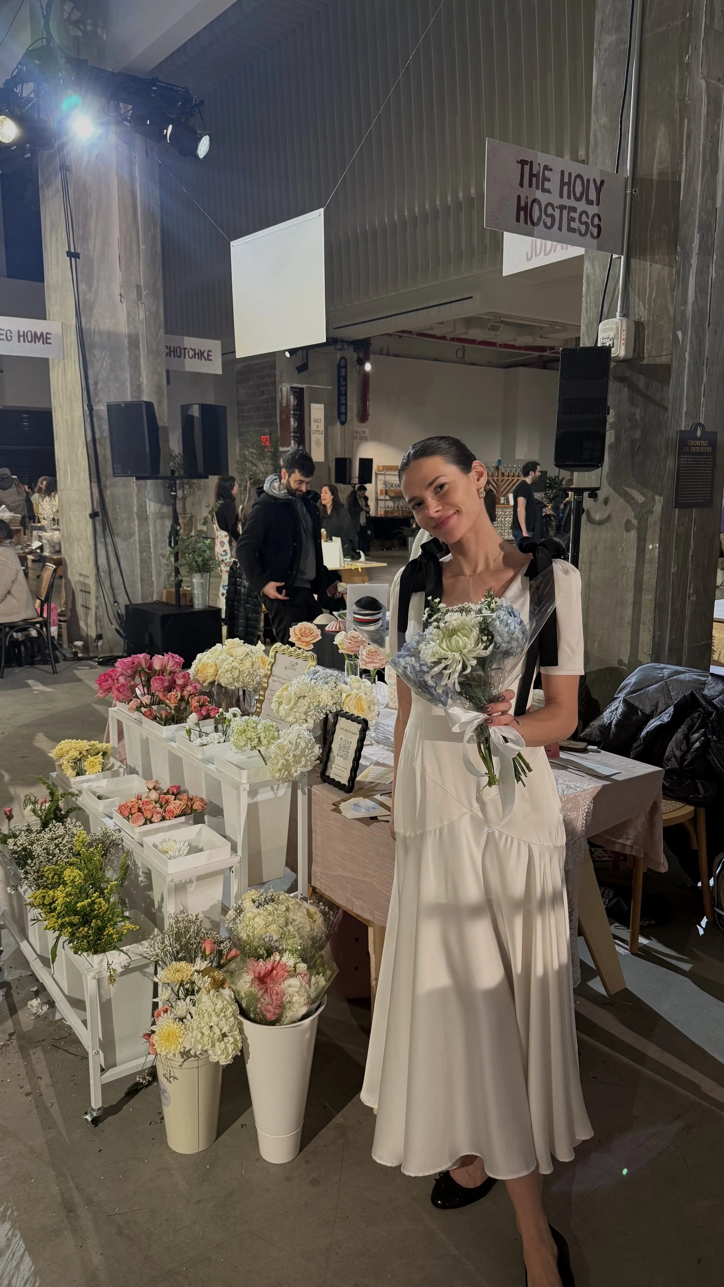 A woman in a white dress standing in front of a flower stall at an indoor market, holding a bouquet of flowers, with various flowers displayed on the stall, and people in the background.