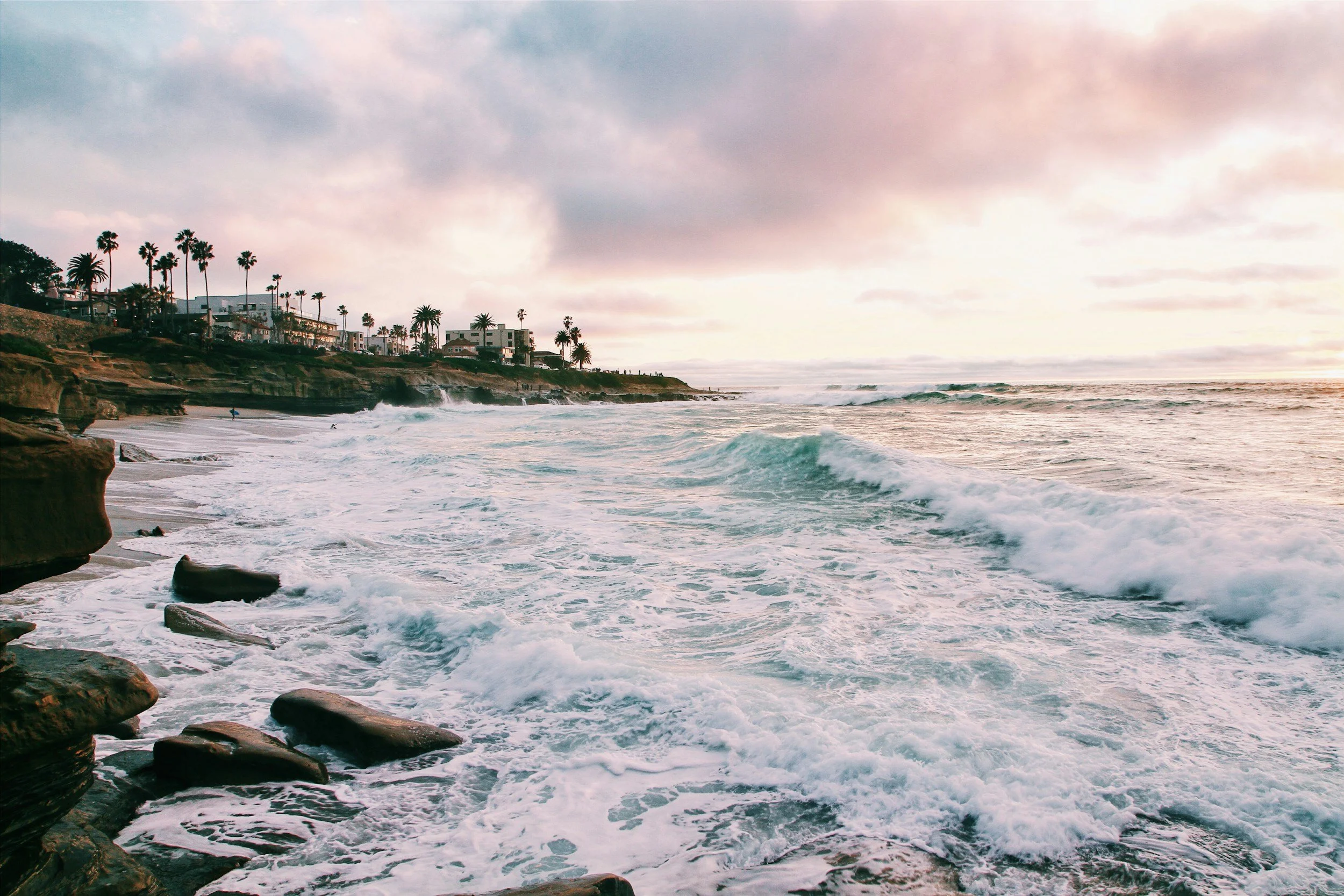 Beach with waves crashing onto rocky shore, houses and palm trees on a hillside, cloudy sky with pink and purple hues.