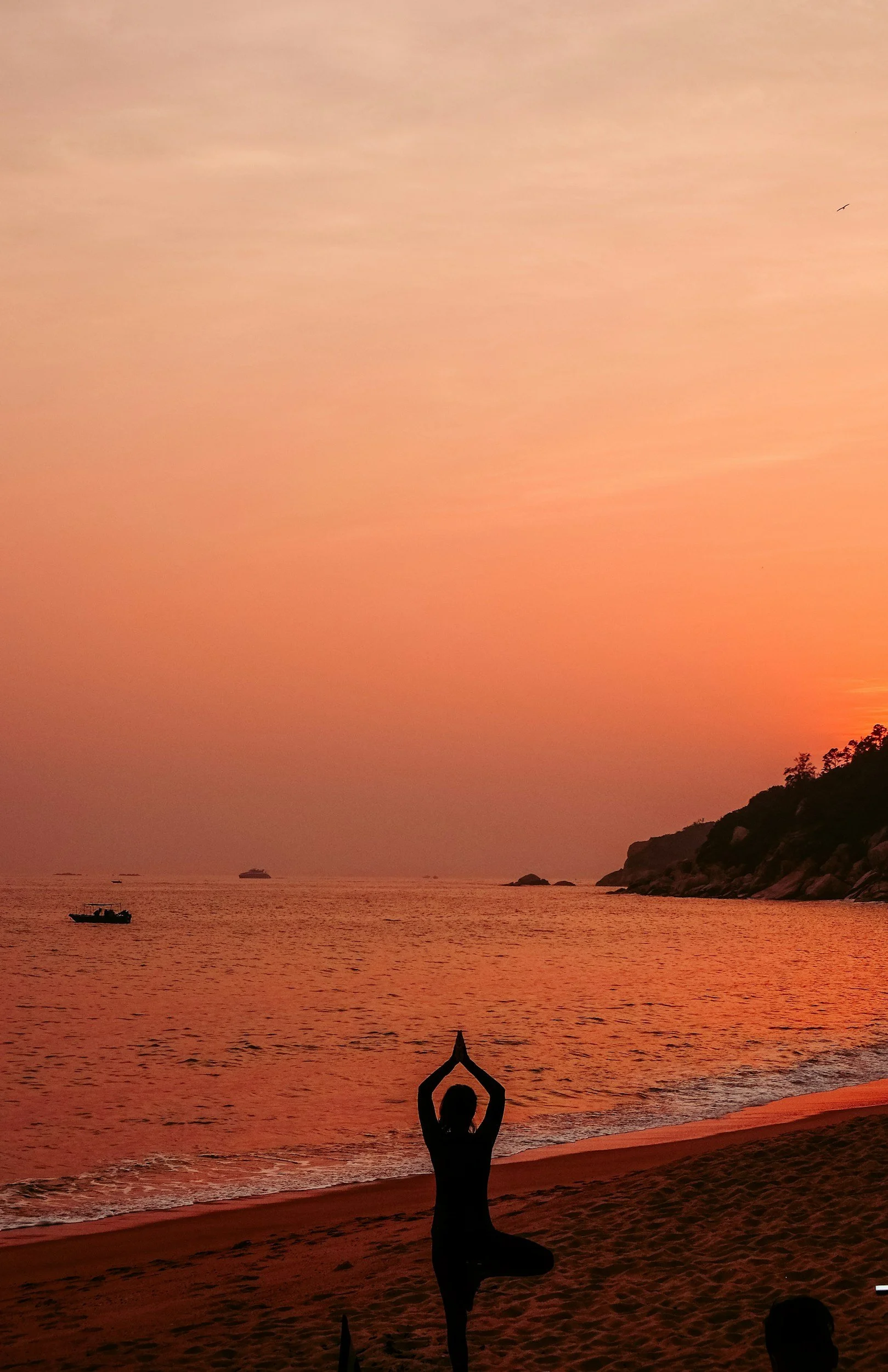 A person practicing yoga in a tree pose on a sandy beach during sunset with an orange-pink sky, boats on the water, and a hillside in the background.