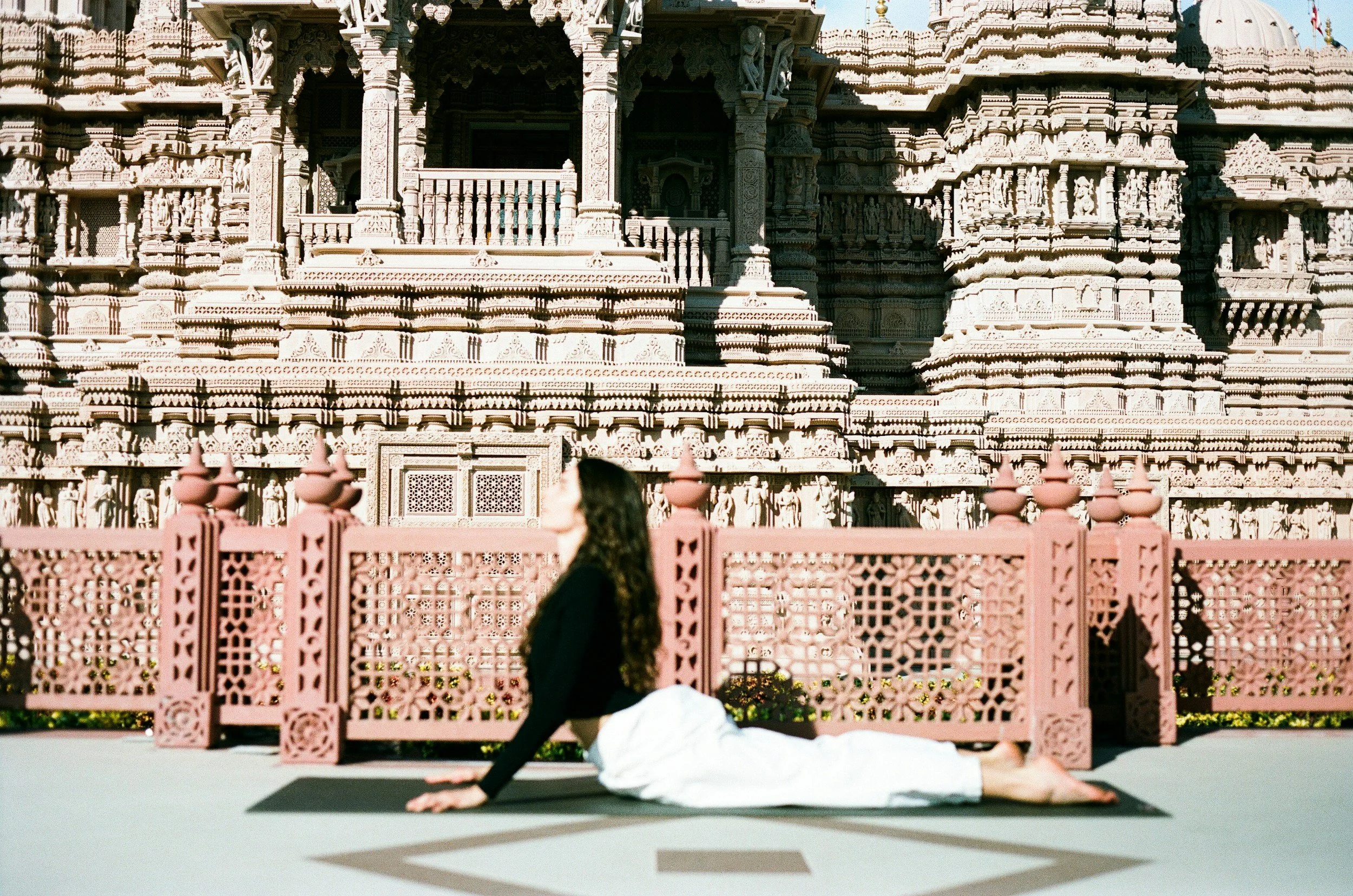 A woman practicing yoga on a mat outdoors in front of a historic temple with intricate carvings and pink fencing.