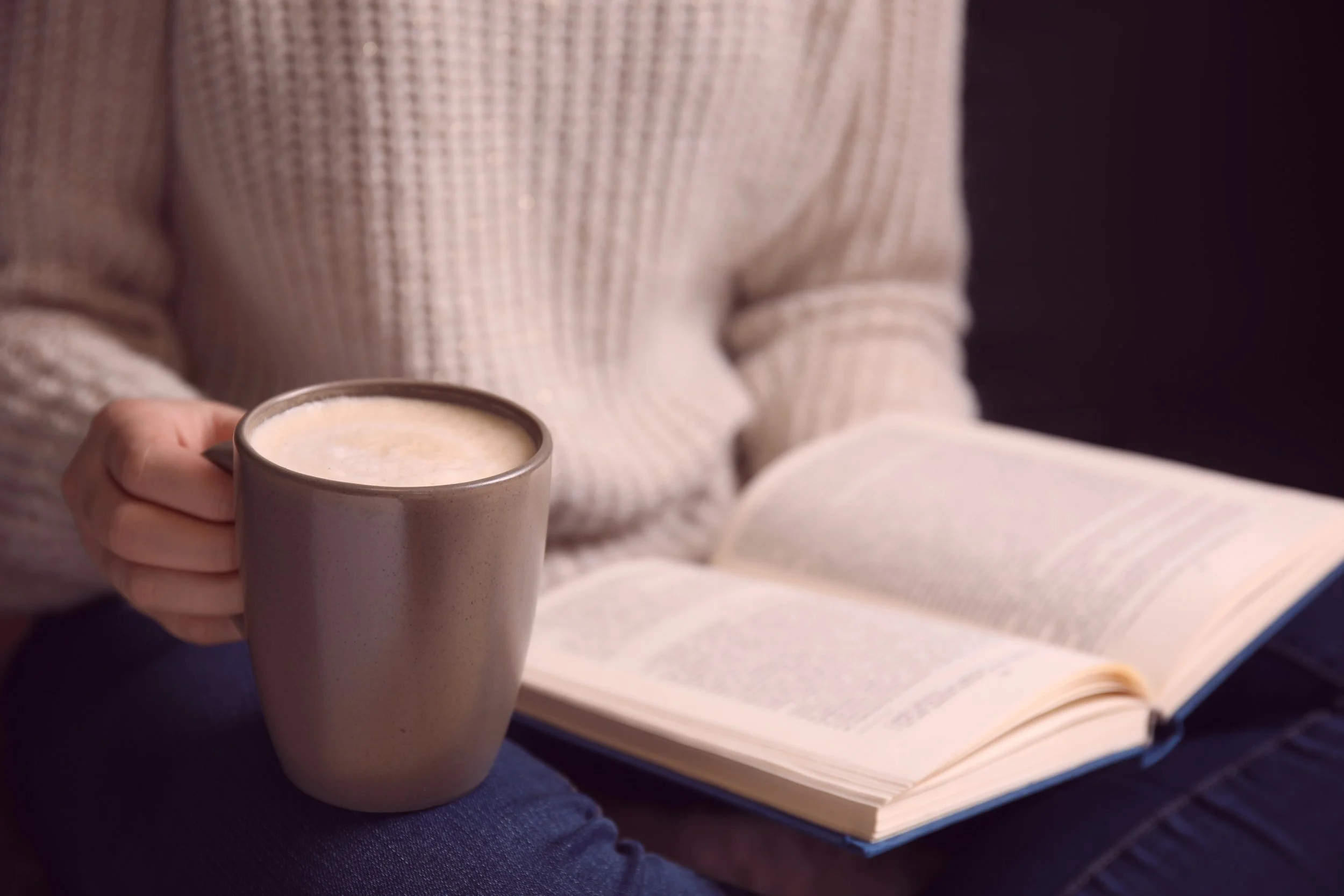 Person in a cream-colored sweater holding a white ceramic mug of coffee or hot chocolate, with an open book on their lap.