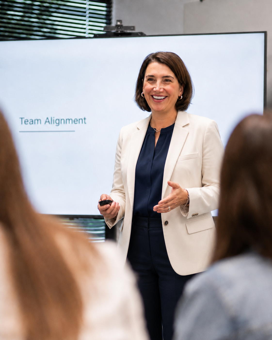A woman wearing a white blazer and dark blue top giving a presentation using a remote in front of a screen that says 'Team Alignment.' She is smiling and gesturing with her hand, with audience members visible in the foreground.