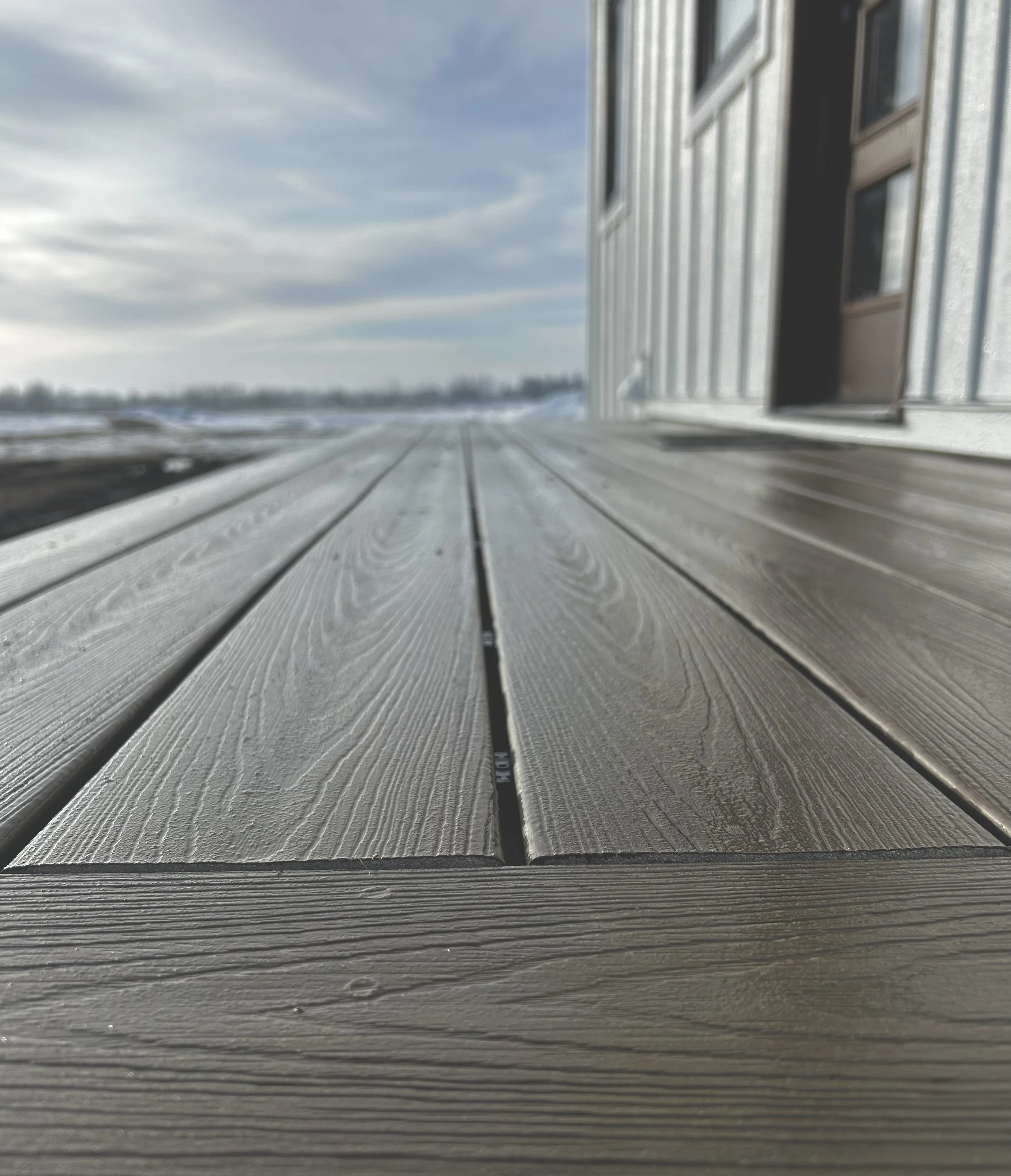 Close-up view of a wooden deck outside a building on a cloudy day, with the building's wall and door visible on the right side and a snowy landscape in the background.