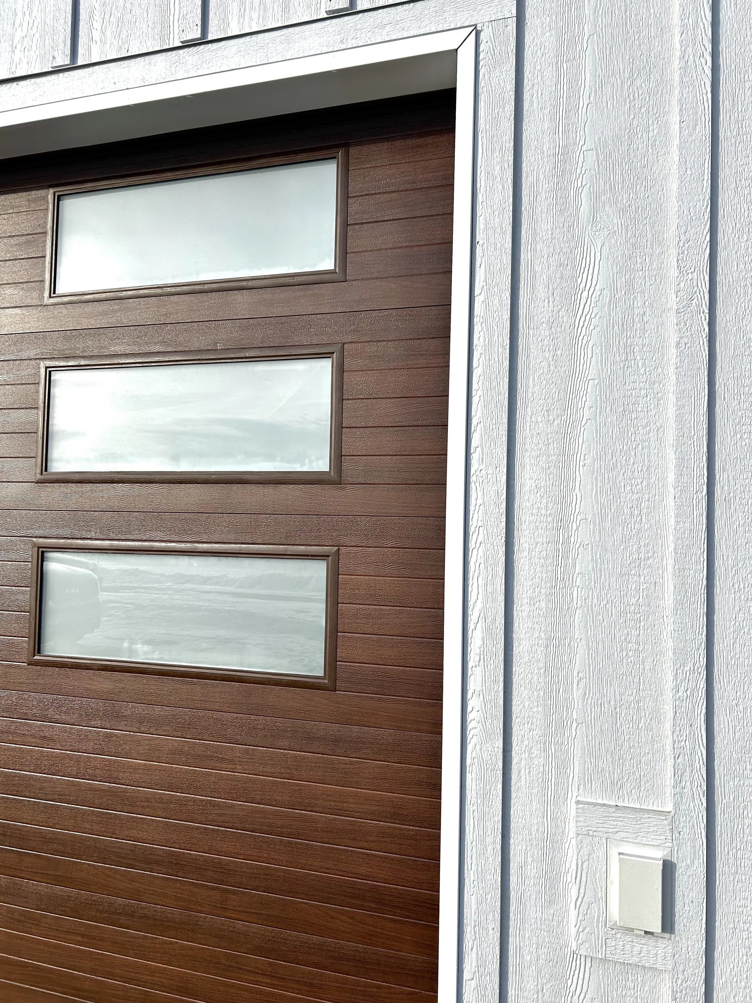 Close-up of a building exterior showing three rectangular windows on a wooden brown wall and a white siding wall with a small utility box.