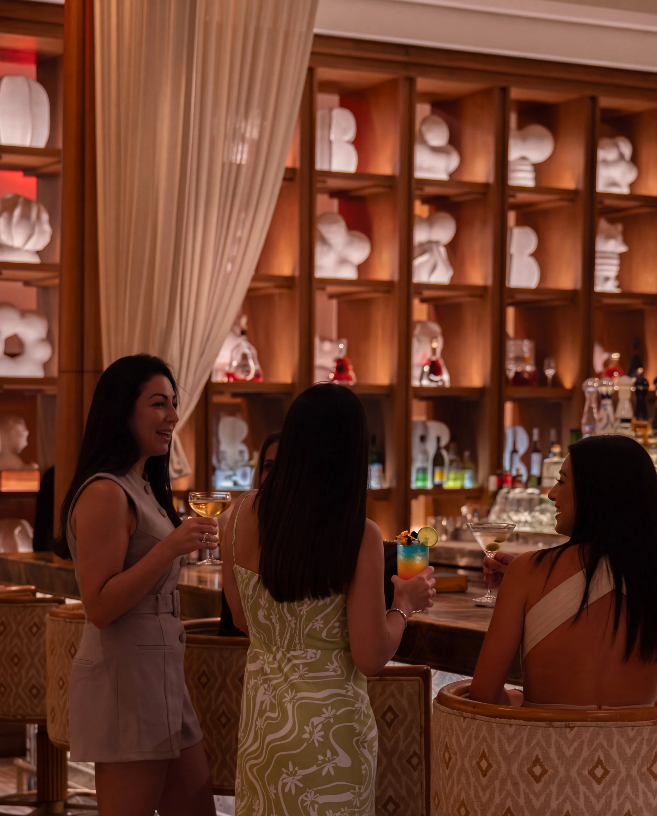 Women having drinks and chatting at a bar in a restaurant with wooden shelving filled with white ceramics and bottles in the background.