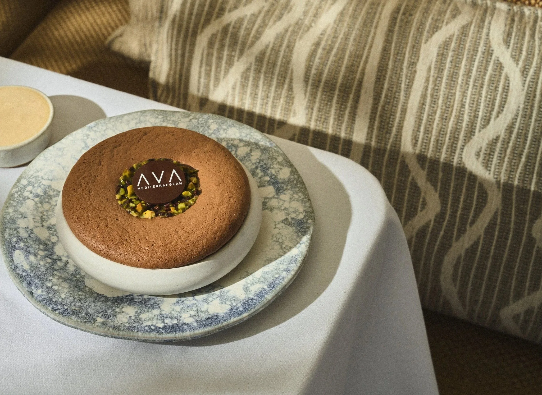 A dessert plate with a round chocolate cake topped with a chocolate disc label reading 'AVA Mediterranean' and sprinkled with chopped pistachios, served with a container of sauce or cream on the side. The plate is on a white table with a textured fabric in the background.