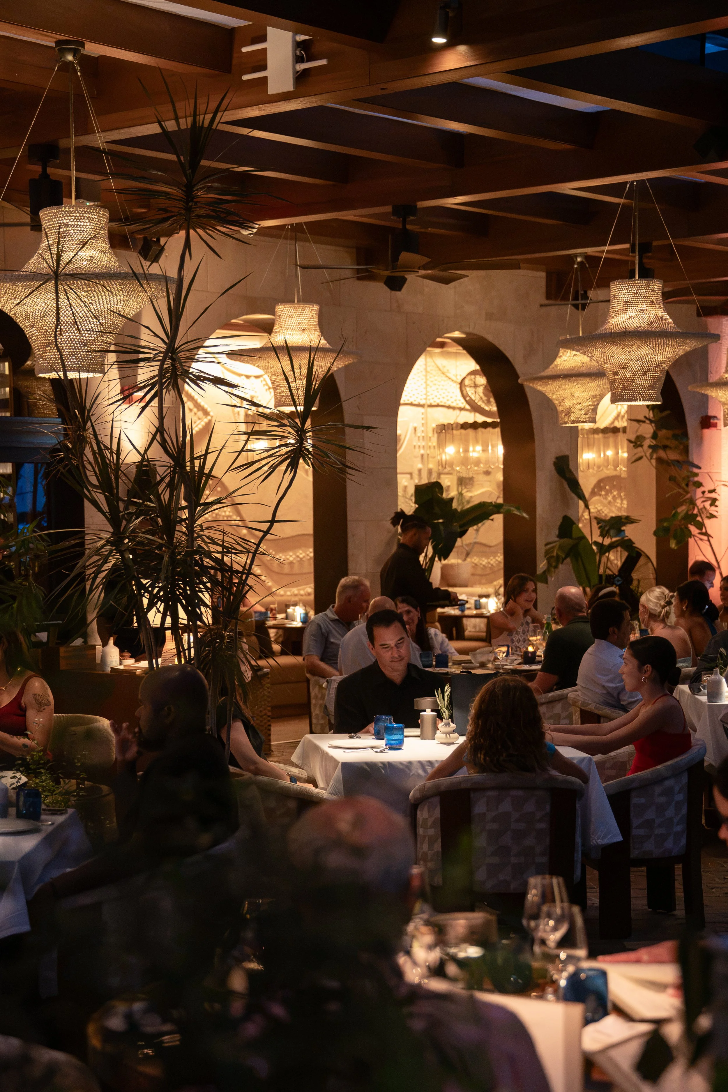 A lively restaurant scene with people dining at tables, warm lighting from woven pendant lamps, and greenery including potted plants, with a wooden ceiling and stone walls in the background.