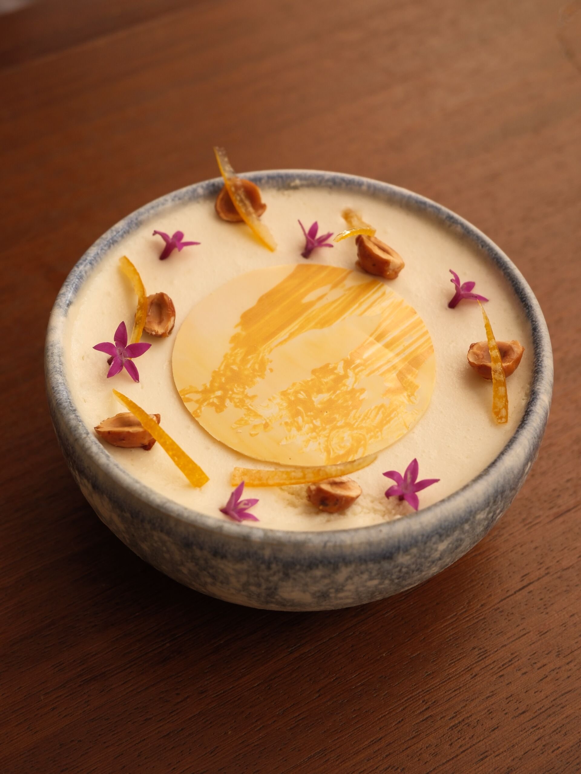 A bowl of creamy dessert garnished with purple flowers, orange zest, and whole nuts on a wooden surface.