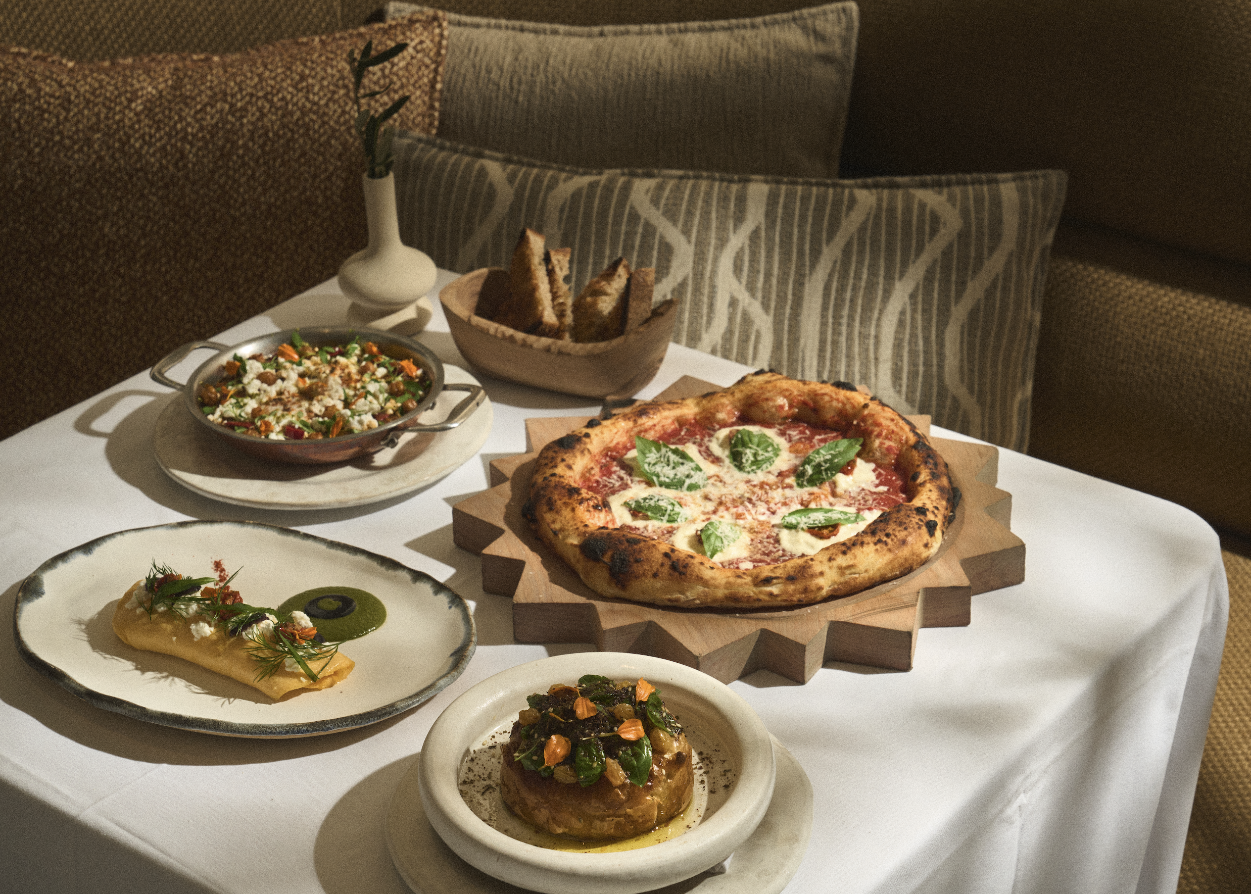 Brunch menu items at AVA Coconut Grove including pizza, an omelet, bread, salad, and a pastry on a table next to a couch and pillows.