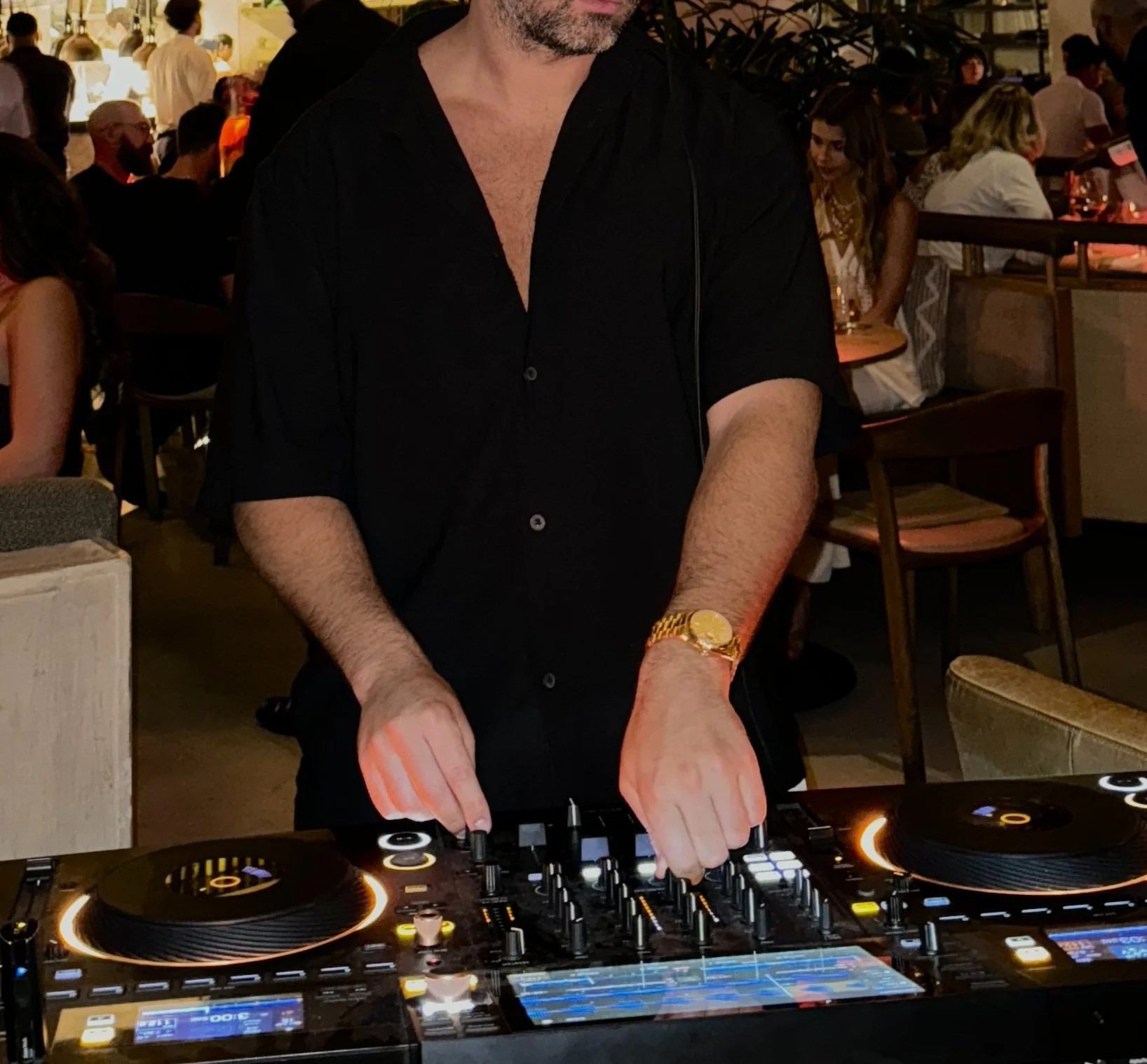A DJ wearing a black shirt with the top buttons undone, a gold watch, and headphones, is mixing music at a DJ console in a lively, dimly lit bar or club, with people socializing in the background.