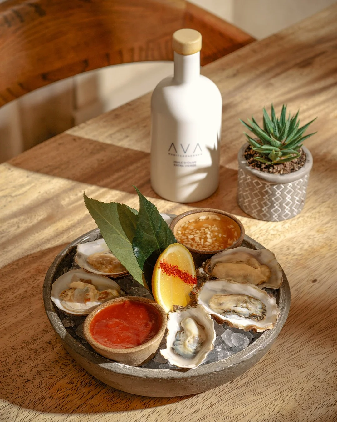 A bowl of fresh oysters on ice with lemon wedge, bay leaves, and dipping sauces on a wooden table, with a white bottle and potted succulent in the background.