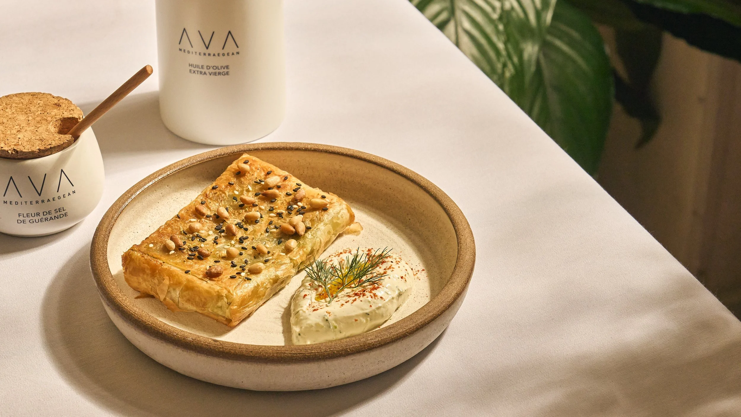 A plate with a rectangular pastry topped with nuts and seeds, accompanied by a dollop of creamy sauce garnished with dill, on a white table with salt and olive oil containers nearby.