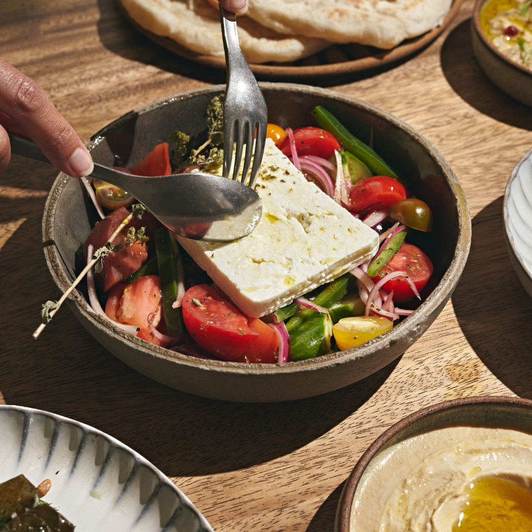 A bowl of fresh salad with tomatoes, cucumbers, red onions, and a large piece of feta cheese on top.