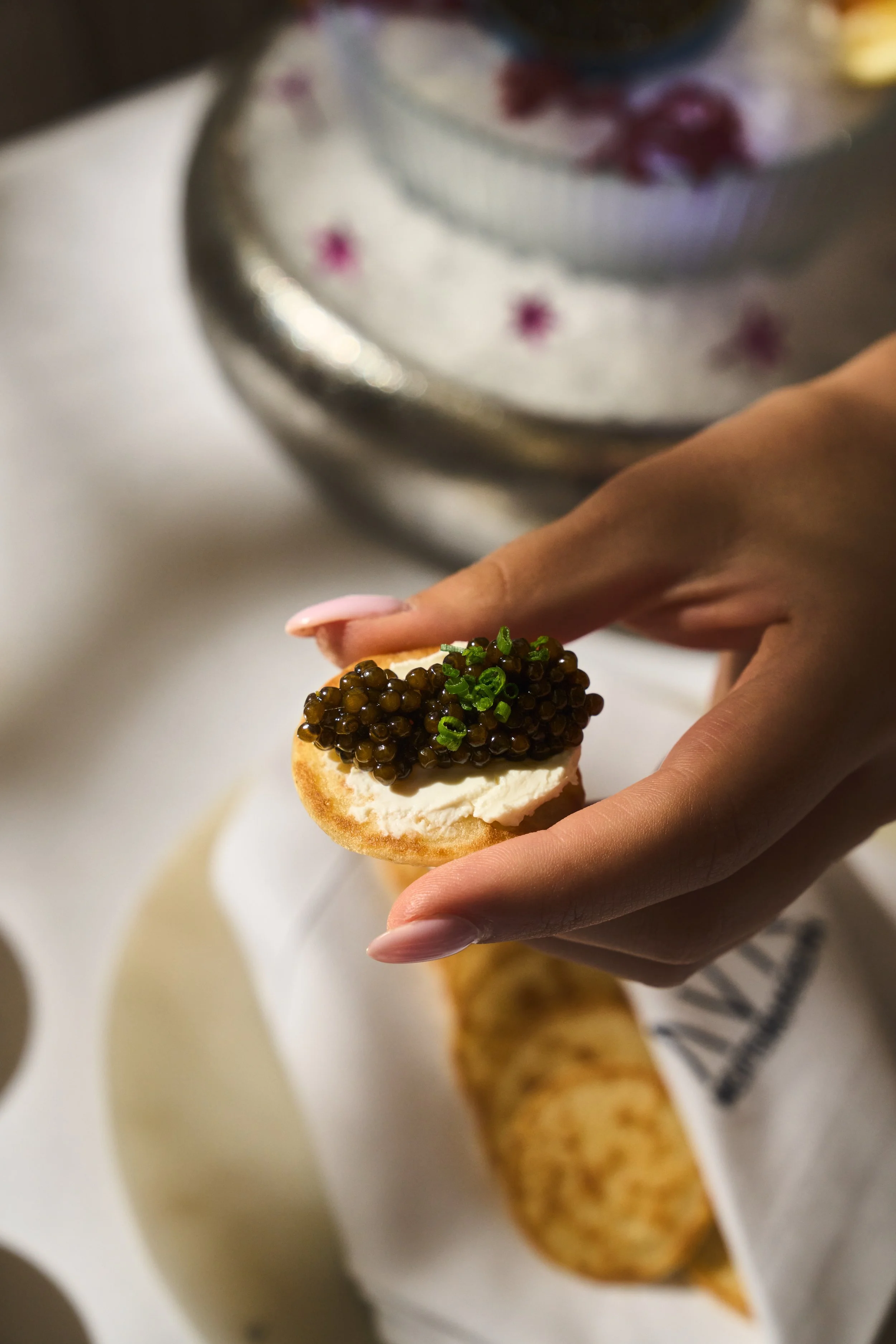 Close-up of a hand holding an appetizer topped with caviar and chopped chives, with a tray of breadsticks in the background.