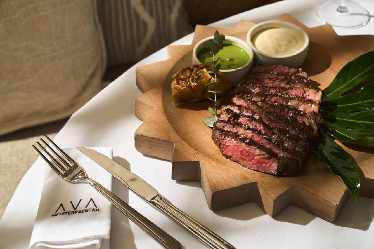 A display of steak on a sun-shaped plate with two sauces, one green colored and beige. There is cutlery and an AVA embroidered napkin besides the dish.