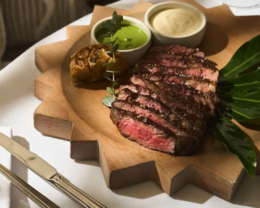 Slices of medium-rare grilled steak served on a wooden platter with dipping sauces, a roasted garlic clove, and garnished with greenery.