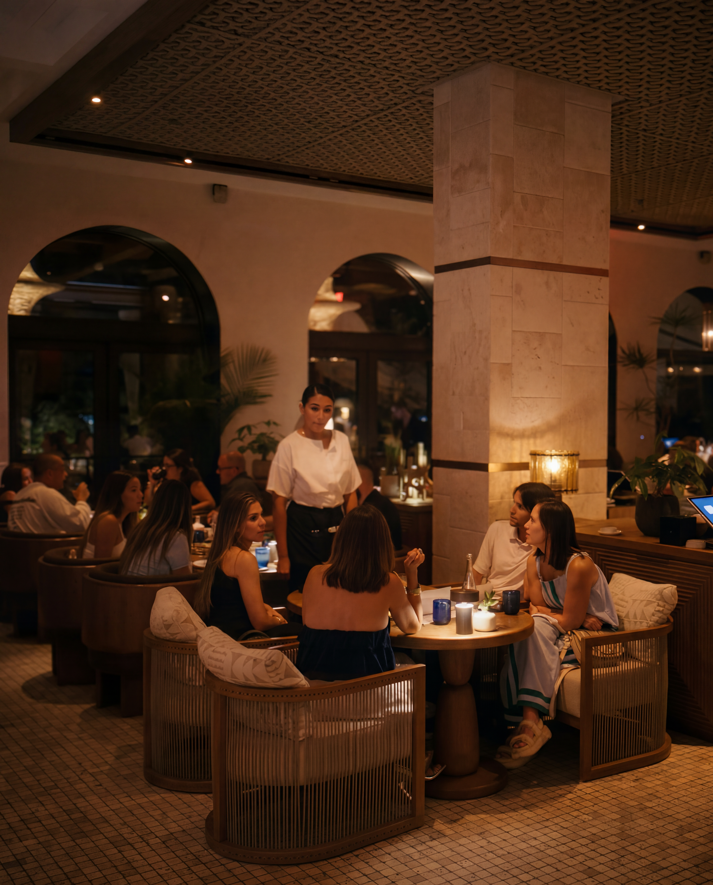 A busy indoor restaurant scene with a group of people dining at wooden tables, led by a waitress in a white top standing near a large marble column. The restaurant has warm lighting, arched windows, a textured ceiling, and a tiled floor.