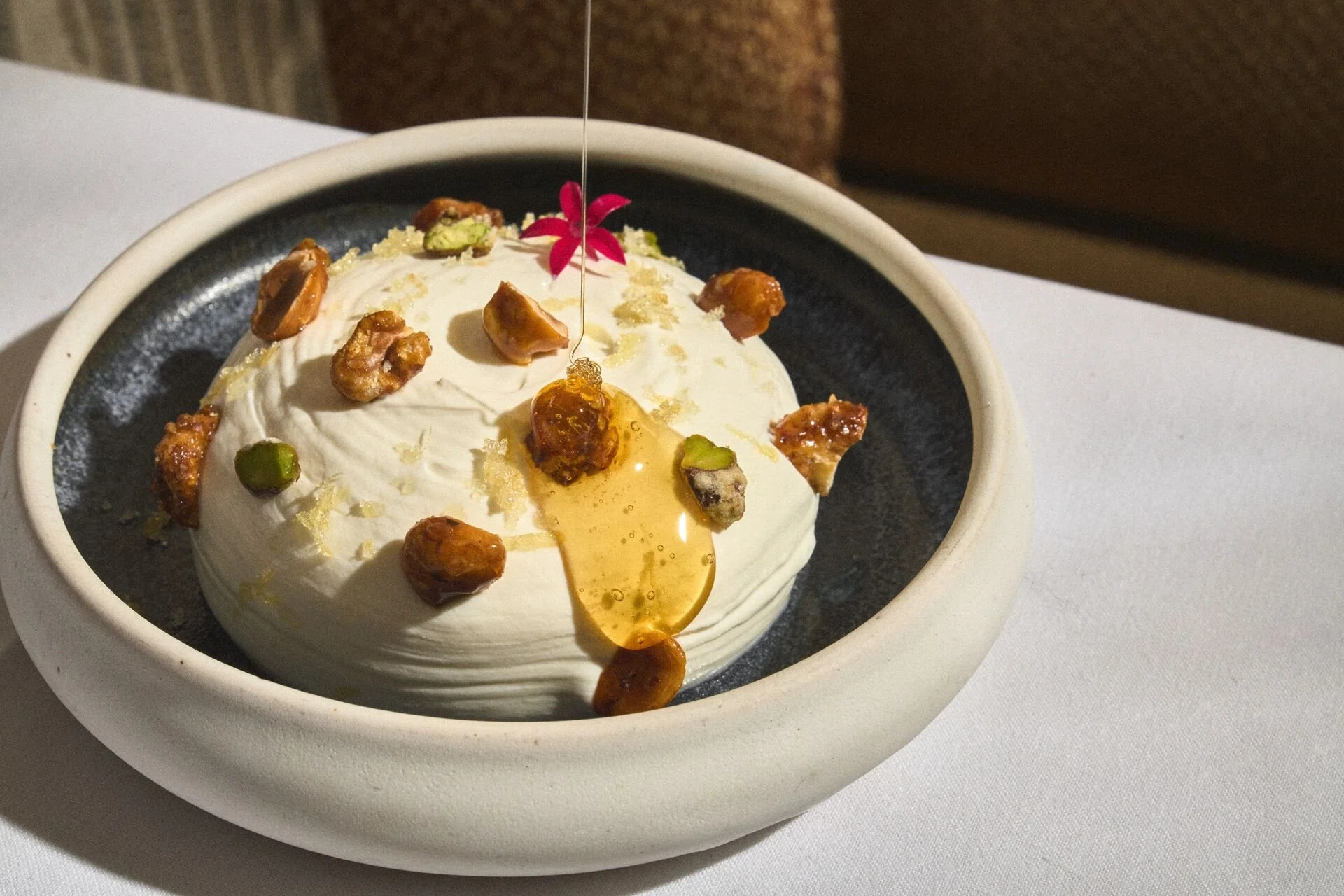 A dessert with a scoop of ice cream topped with a pink flower, golden syrup, chopped nuts, and pistachios, served in a black bowl with a white exterior, on a white table.