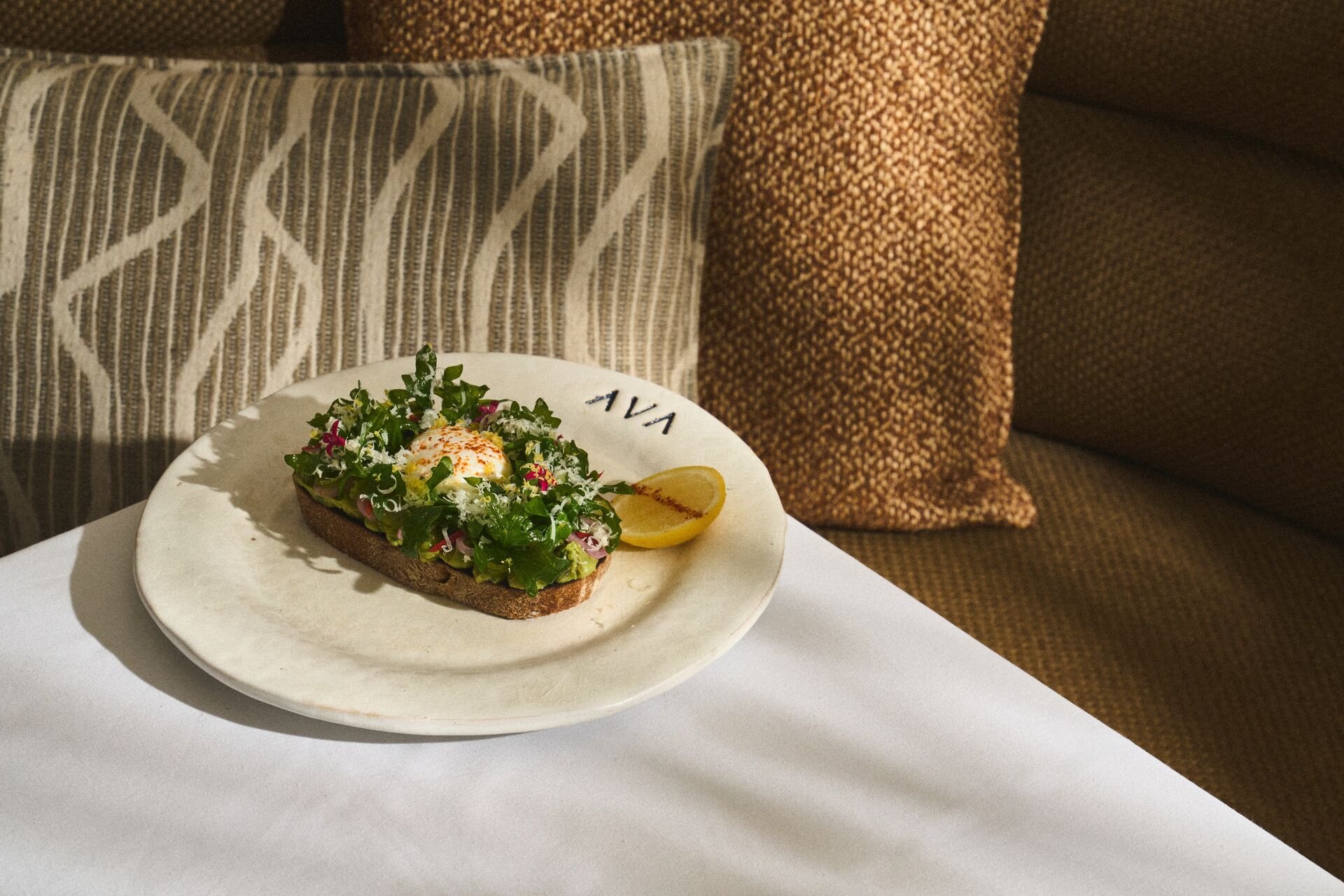A plate of avocado toast with fresh herbs, radishes, and a poached egg, garnished with paprika and a lemon wedge, on a white table in front of patterned cushions.