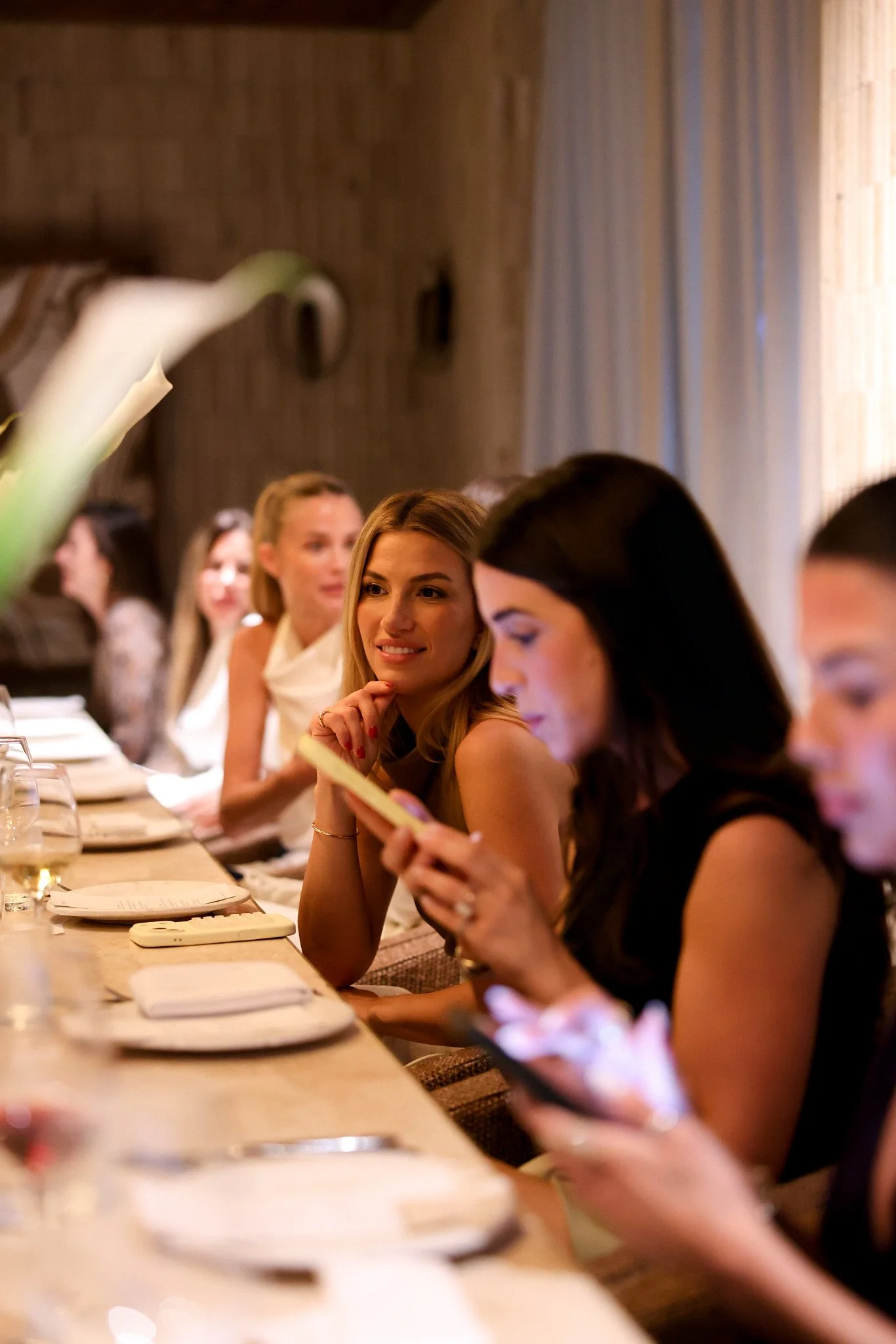 A group of women sitting at a dining table during a gathering or dinner, with some women looking at menus or phones and engaging in conversation, in a warmly lit setting with a wood-paneled wall in the background.
