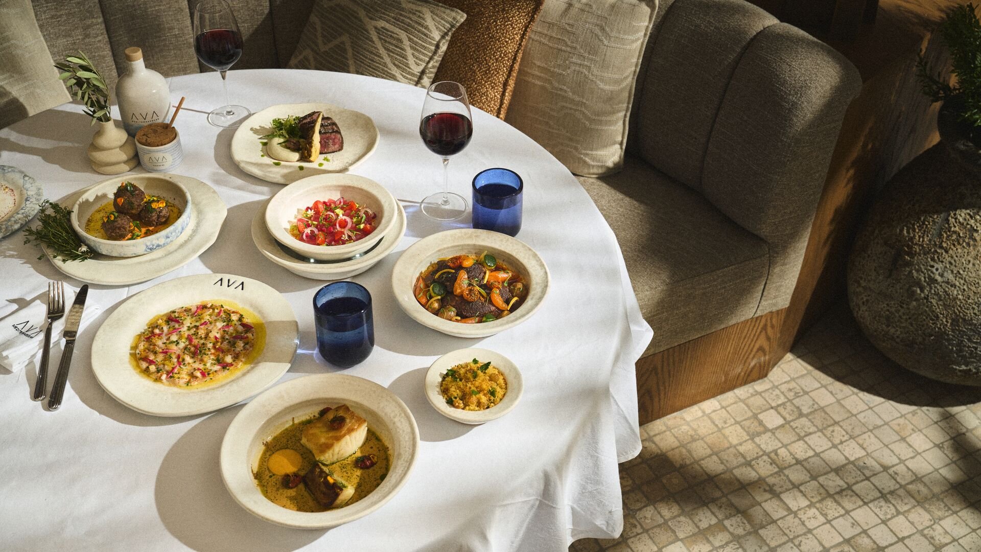 A table set with a variety of dishes including salads, meats, and a fish entrée. There are two glasses of red wine, several blue water glasses, and utensils on a white tablecloth in a cozy dining area with upholstered chairs and decorative vases.