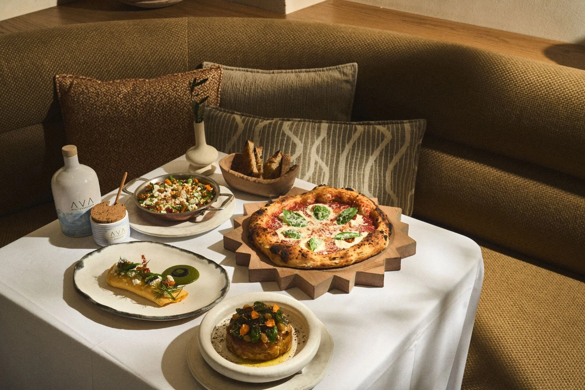 A table with various dishes including a pepperoni pizza on a wooden tray, a salad, a stuffed pastry, and other plated foods, set against a background of cushioned seating with patterned pillows.