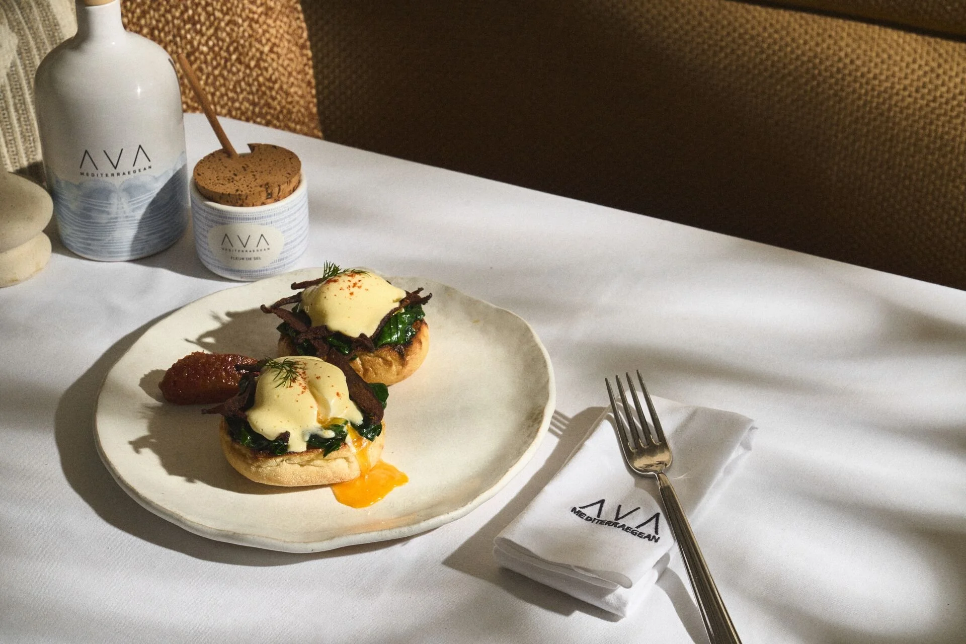 Plate with two poached eggs on bread, topped with greens and served with a dollop of sauce, on a white tablecloth with a fork and a napkin nearby.