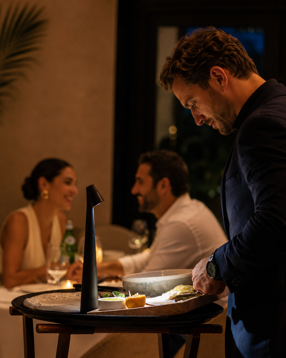 A man in a dark suit serving food at a restaurant table while a couple in the background is happily talking and smiling.
