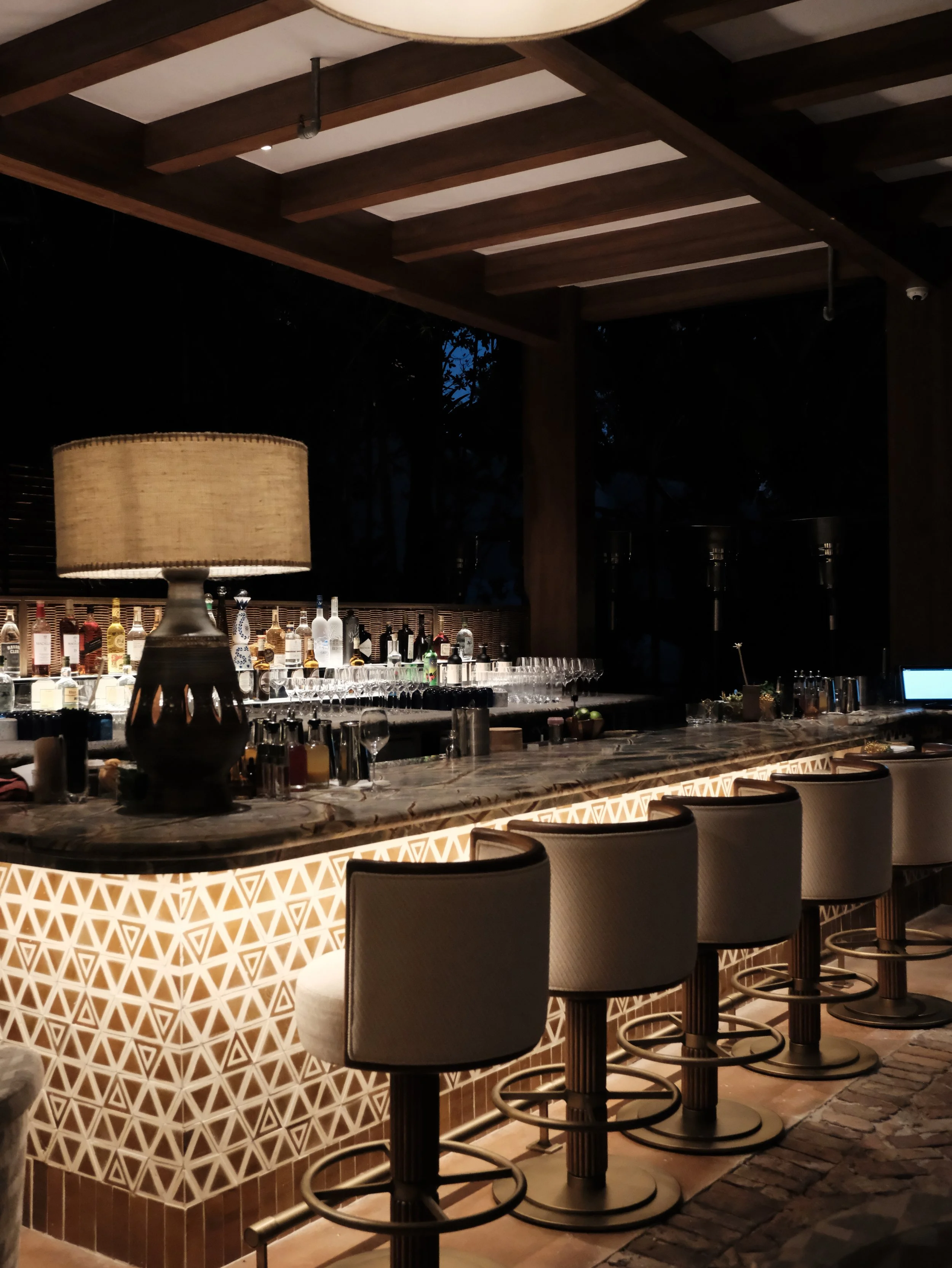 A bar area with a patterned front, high chairs lined up, a marble countertop, liquor bottles behind the bar, wine glasses on the counter, and a large lamp with a beige shade on the left side, under a wooden ceiling with beams.