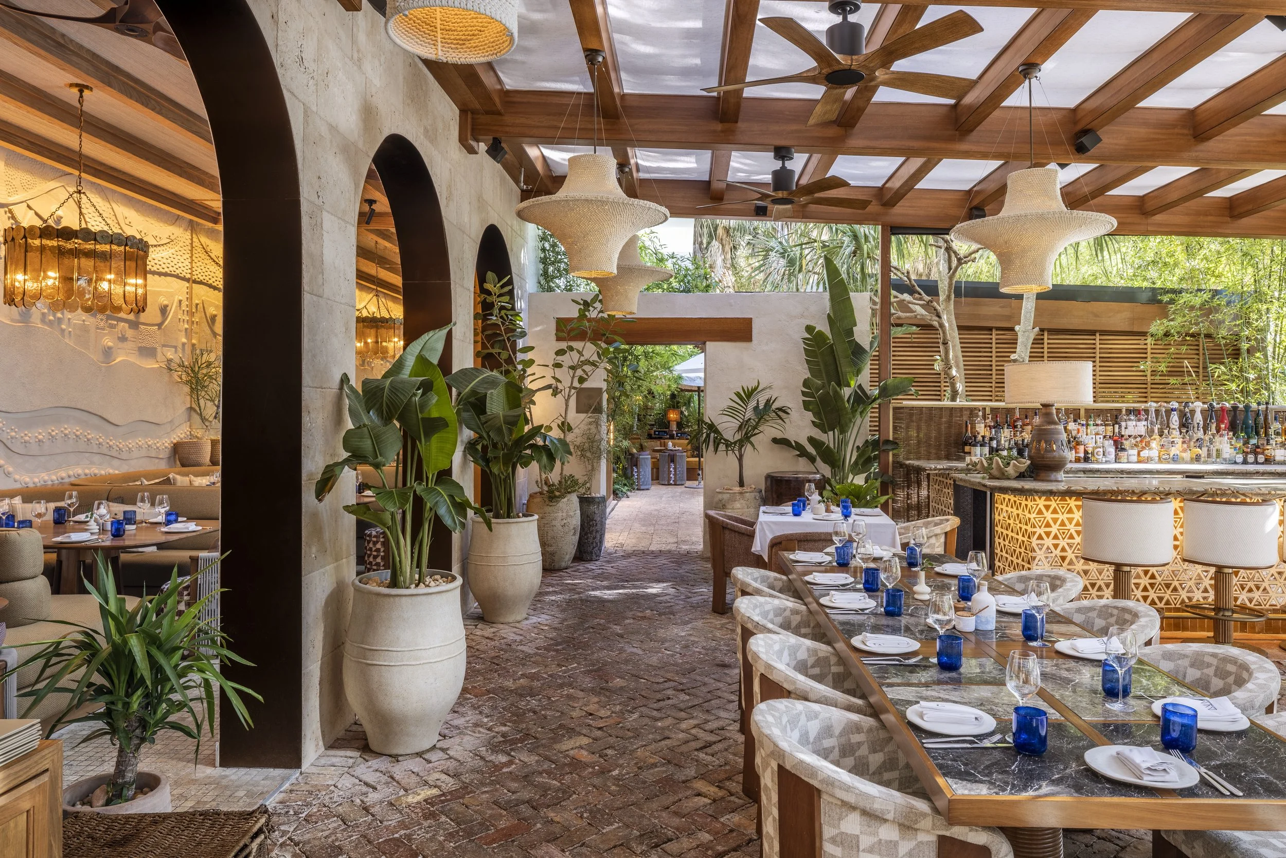 Indoor restaurant with brick flooring, large green plants, white curtains, tables set with white plates, glasses, and blue cups, and hanging woven light fixtures, with a bar area in the background and outdoor greenery visible through an open entrance.