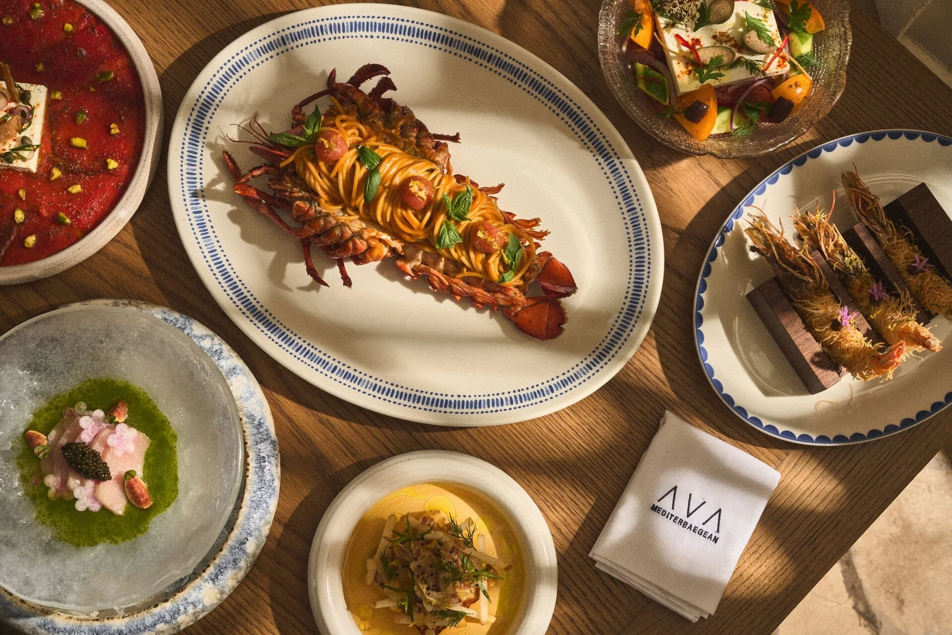 Various gourmet dishes on a wooden table, including a lobster tail with spaghetti, a tomato salad, a seafood platter, and other colorful appetizers.