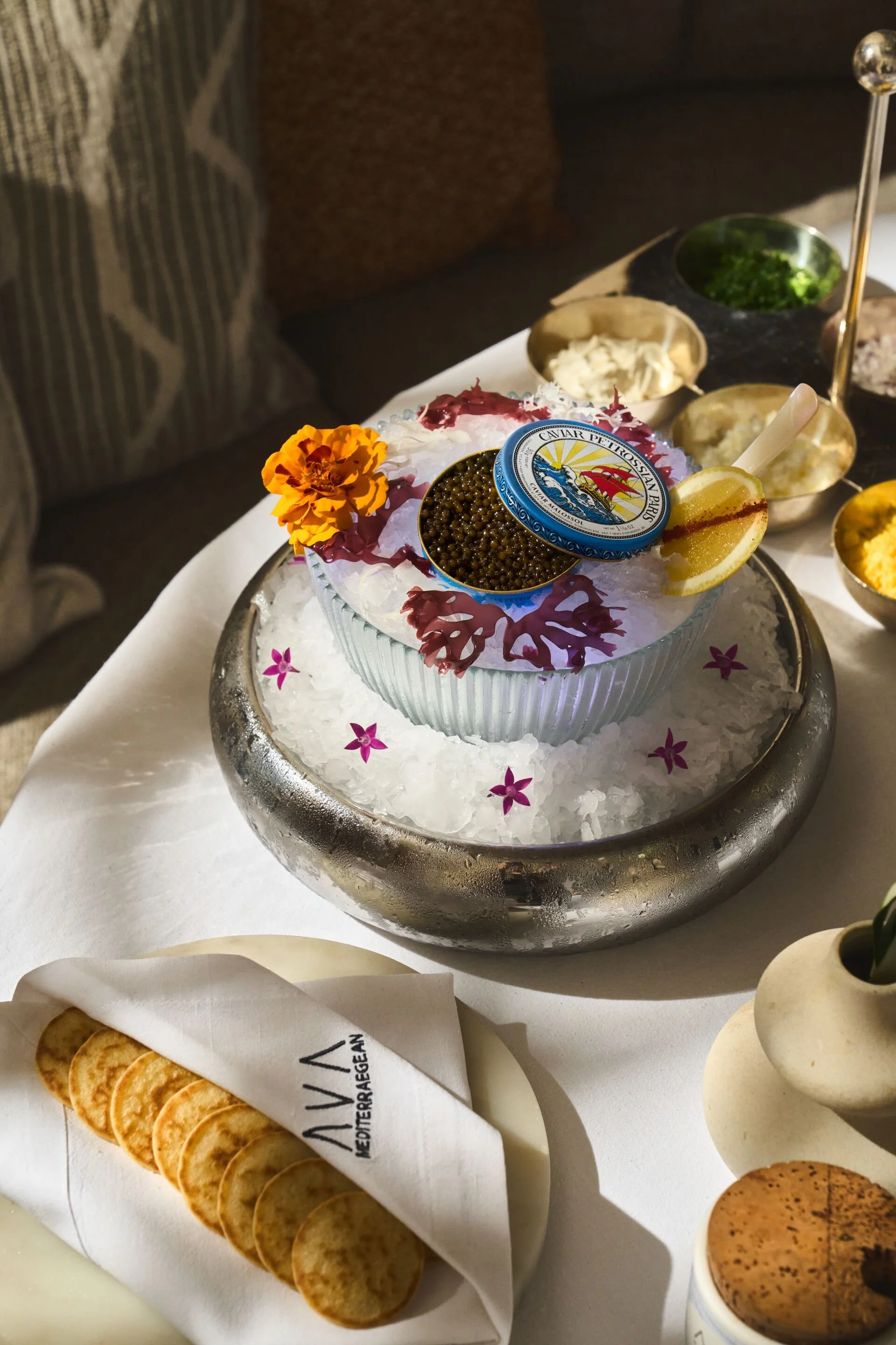 A platter of caviar with a lemon wedge, flowers, and decorative leaves on ice, accompanied by various cheeses, crackers, and spreads in bowls on a table.