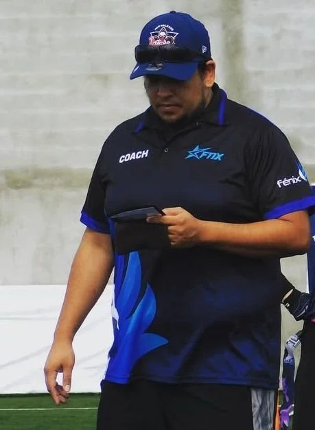 A man wearing a blue and black sports uniform with a logo and the word "COACH" on it, holding a phone, standing outdoors.