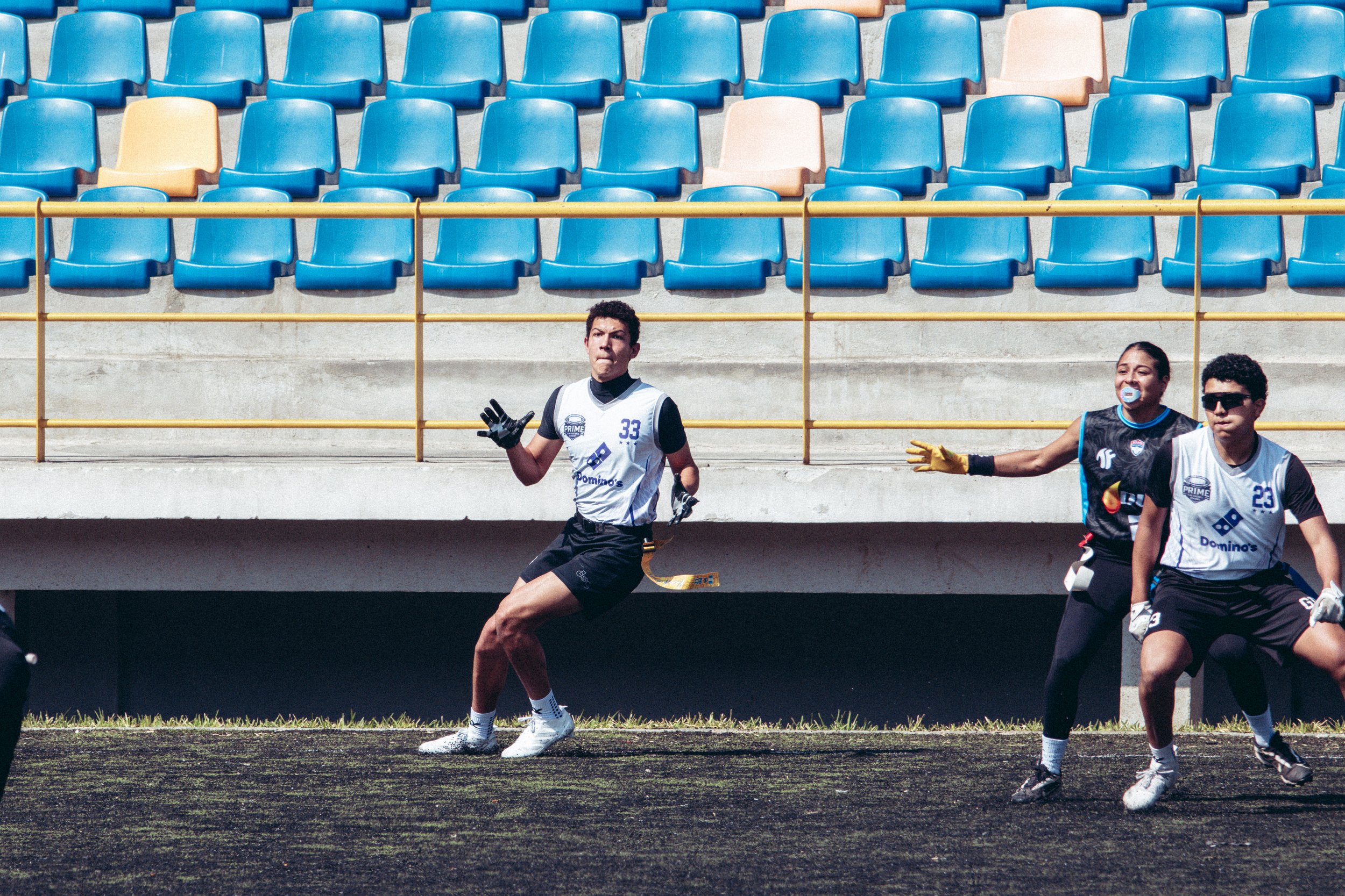 Soccer players during a game, with three players in focus on the field, one in black and two in white jerseys, set against empty blue and peach-colored stadium seats.