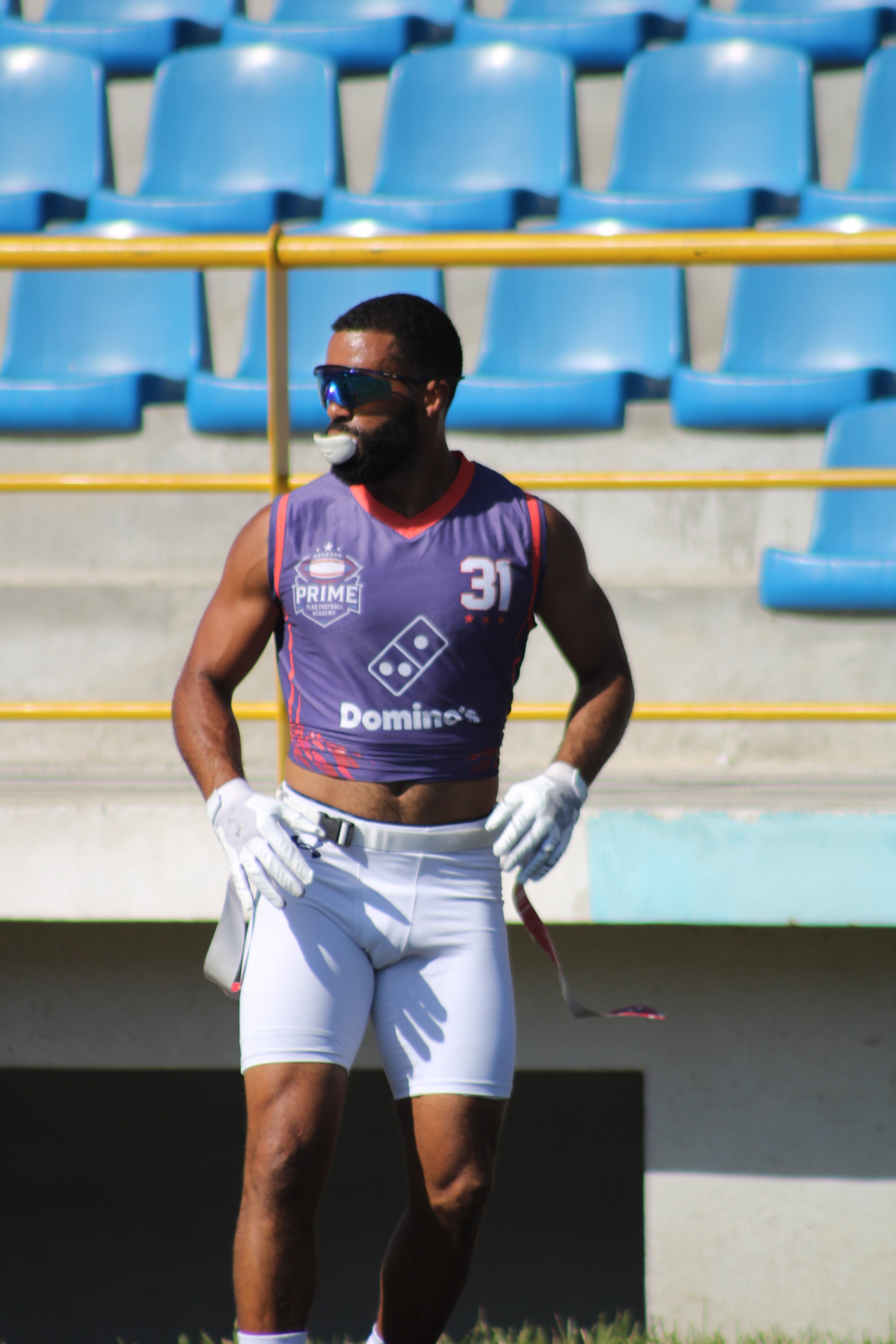 A man in athletic gear standing on a sports field, wearing sunglasses, gloves, and a sports jersey with the logo of Domino's Pizza and the number 31.