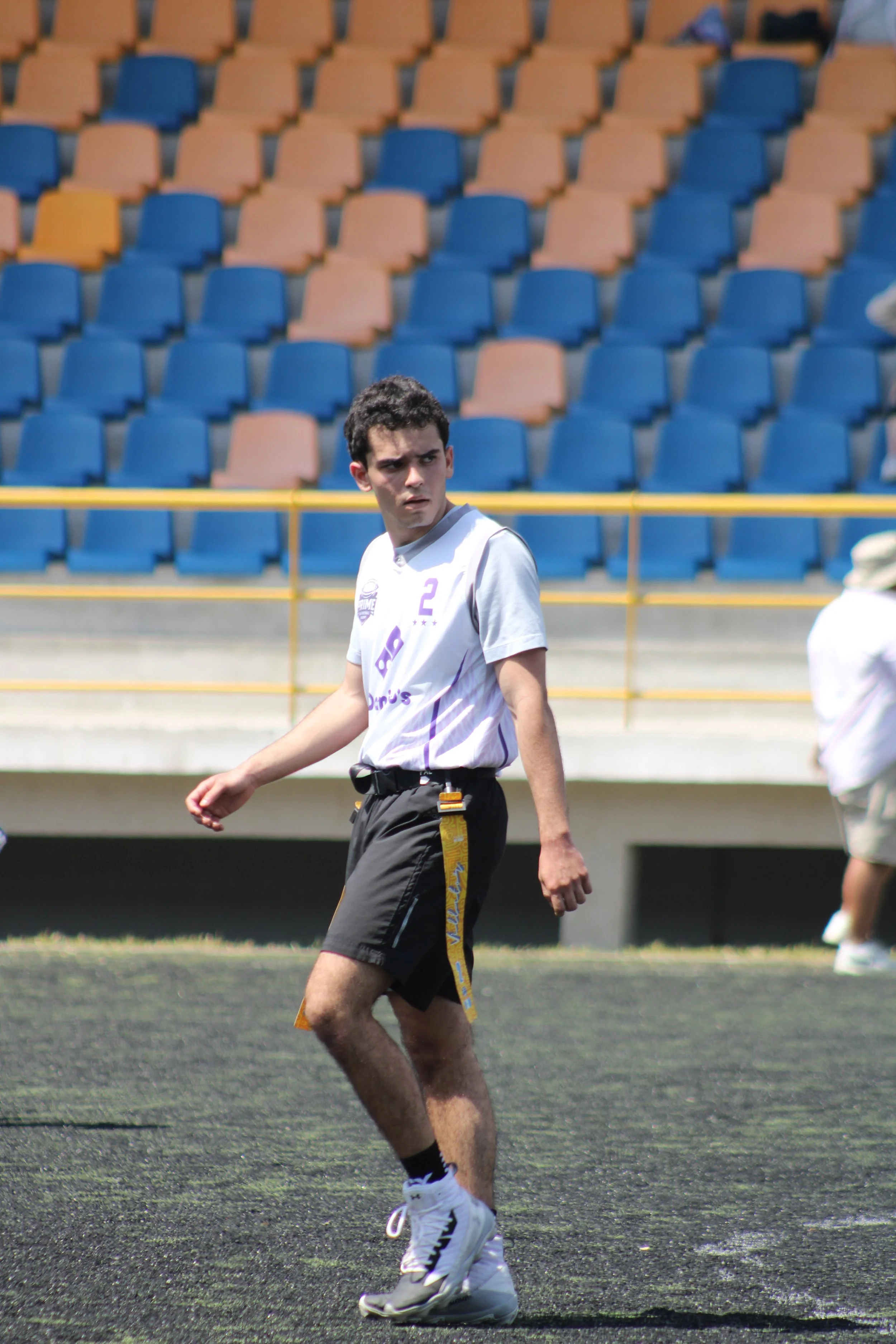Young male athlete in a white sports jersey, black shorts, and white sneakers on a sports field, surrounded by empty colorful stadium seats.
