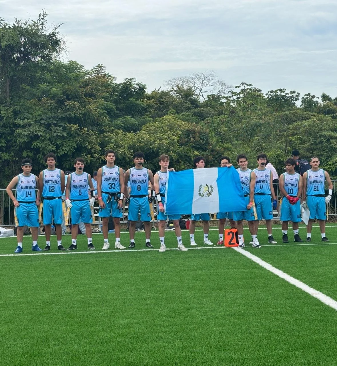 A group of young male basketball players in light blue and white uniforms standing on a sports field, holding the Guatemalan flag. They are wearing black microphones on their faces. The background features green trees and a cloudy sky.