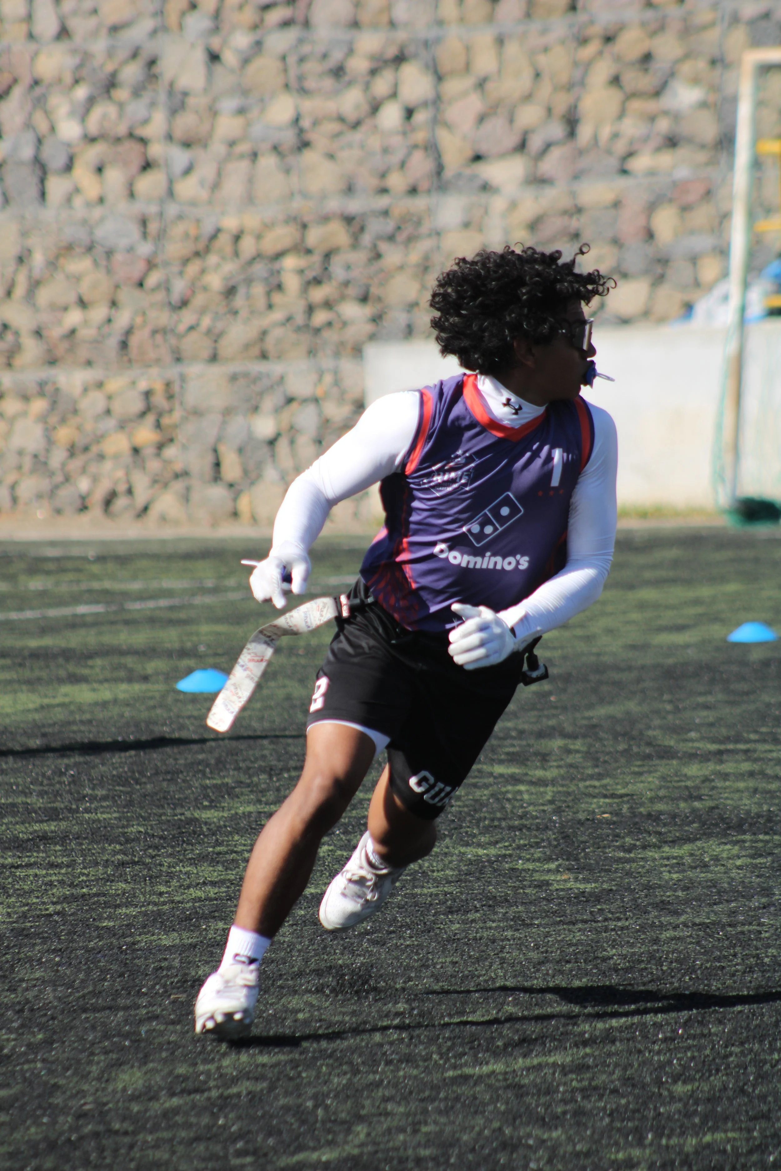 A football player in a dark blue and red jersey, black shorts, and white cleats running on a field during practice or a game.
