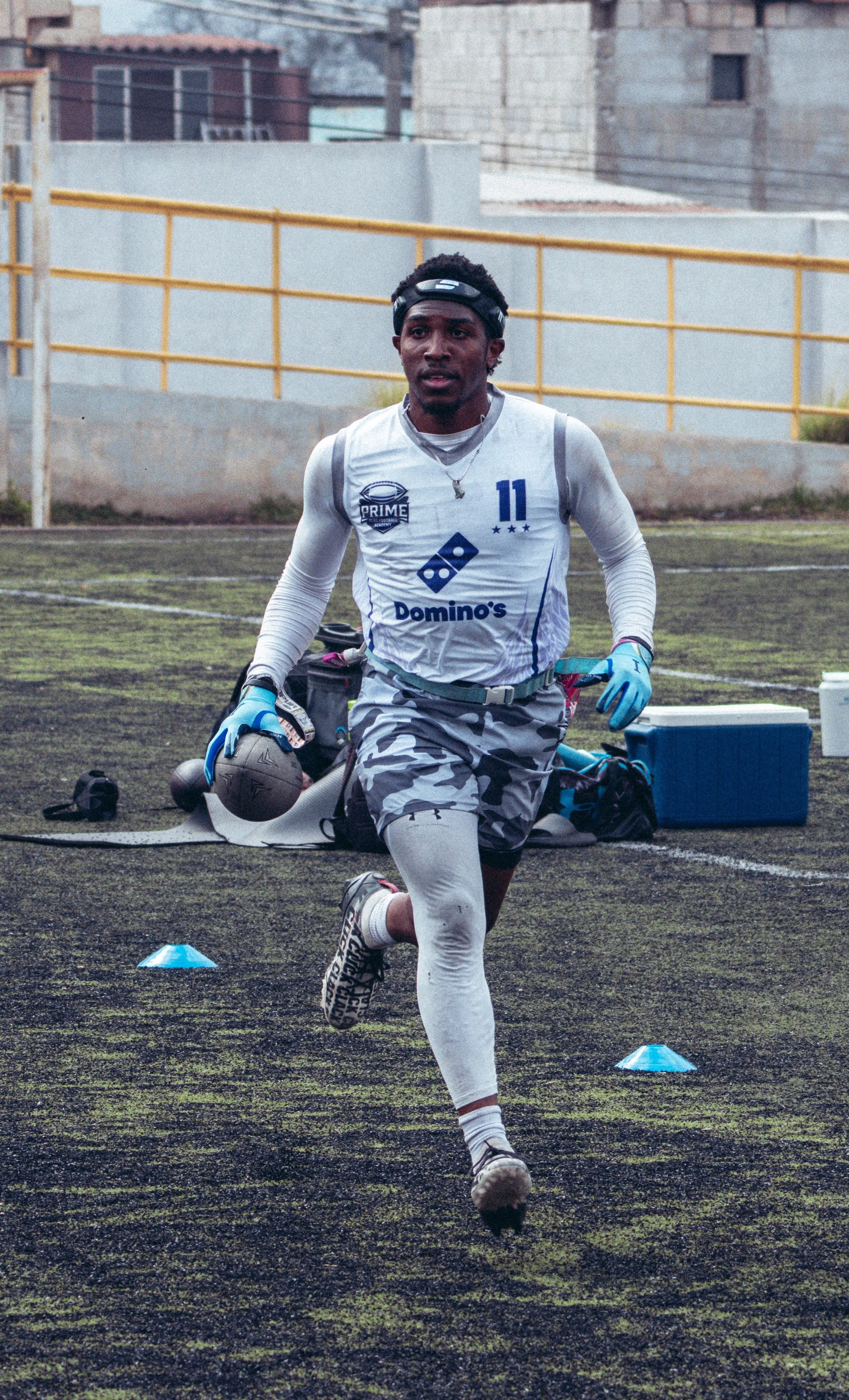 An athletic man wearing a white sports jersey with the number 11, camouflage shorts, white compression leggings, and blue gloves is running on a field while carrying a football. There are training cones and gear in the background.