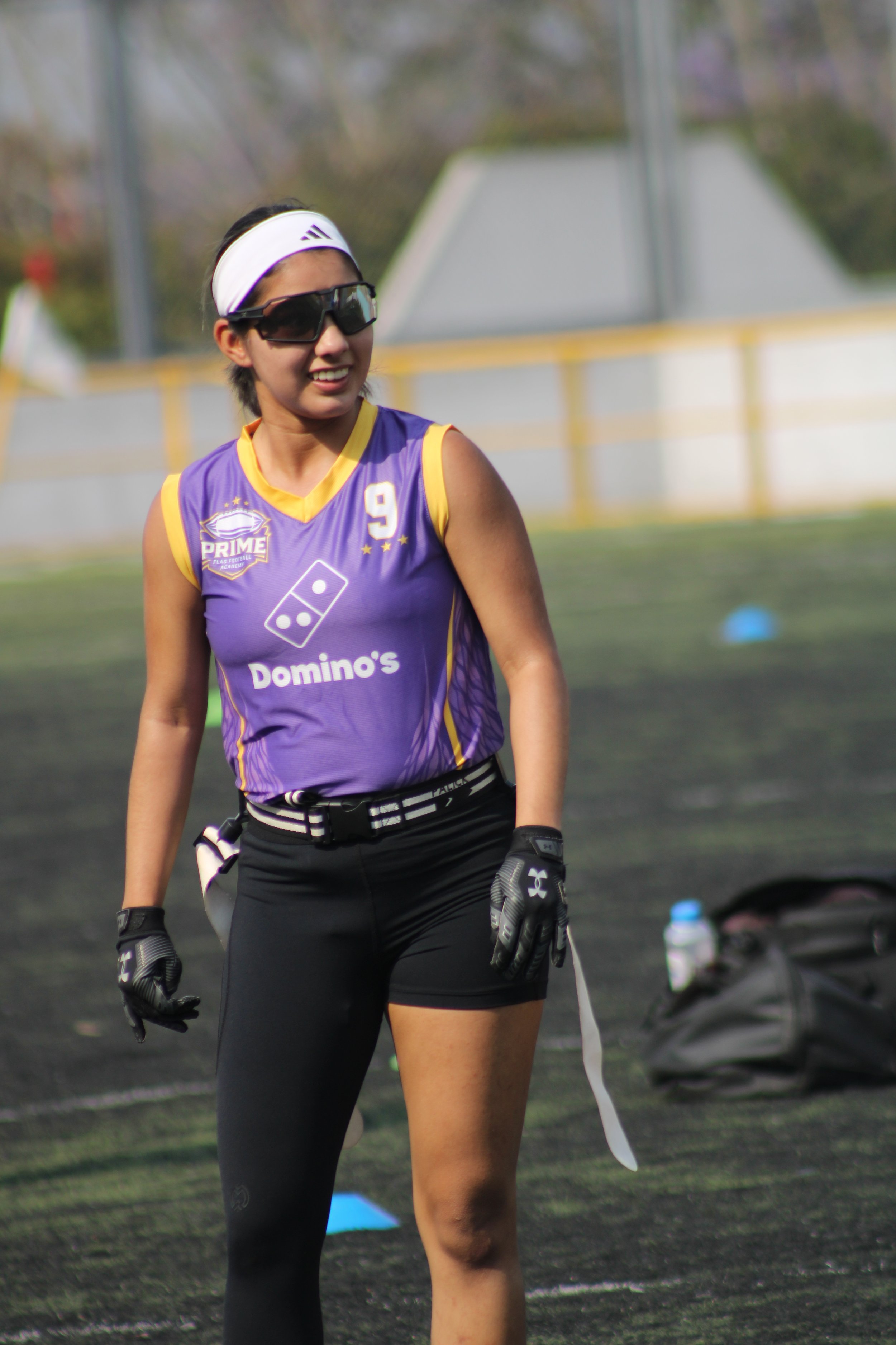 A female football player wearing a purple and yellow uniform with the number 9, sunglasses, a white visor, and gloves, standing on a football field.