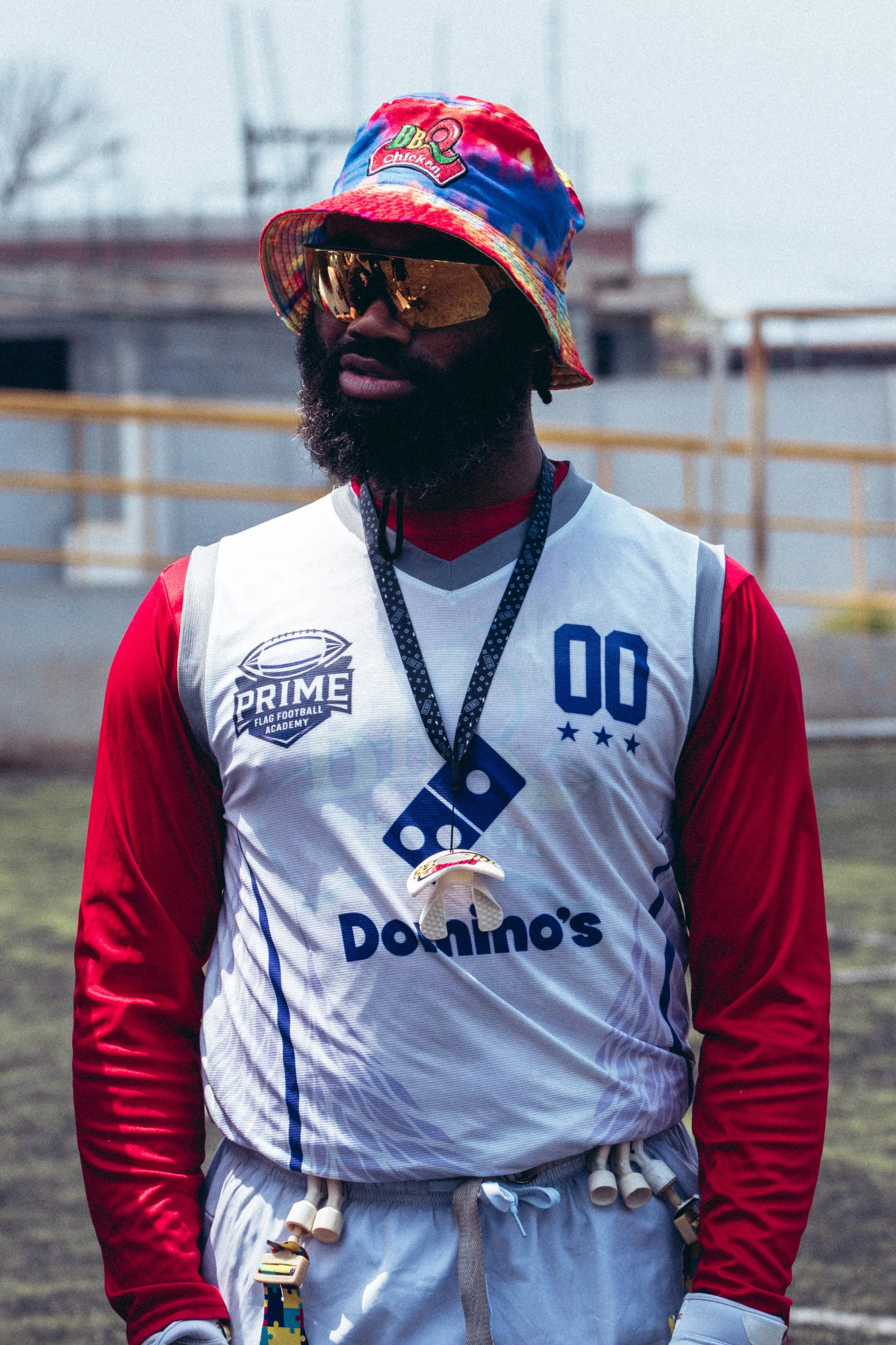 Man wearing colorful bucket hat, sunglasses, and a football jersey with a Domino's sponsorship, standing outdoors on a sports field.